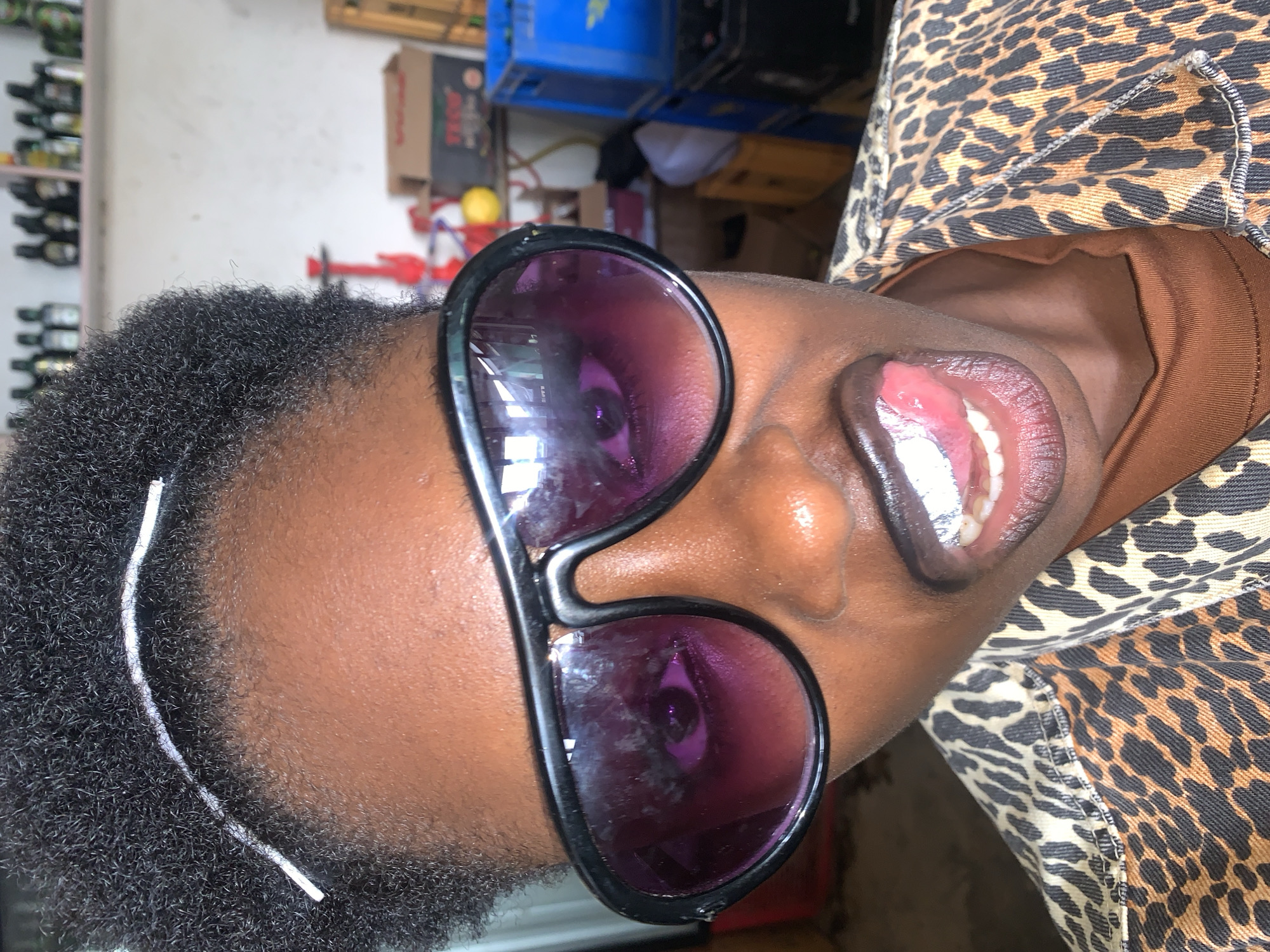 The media file is a close-up, rotated image of a person wearing large, dark-rimmed sunglasses with purple lenses. The person has dark skin and short, dark, curly hair, with a white hair tie visible around the side of their head. Their mouth is open, revealing teeth and a pink tongue. They are wearing a jacket with a leopard print pattern over a brown garment. The background appears to be indoors, with shelves and boxes visible, suggesting a storage or retail environment. Some of the items in the background include blue plastic containers, cardboard boxes, and what looks like bottles on shelves. The lighting suggests it could be daytime, but no specific weather conditions are visible. There is no discernible text in the image. The context provided indicates the location is New Stadium, Nigeria.