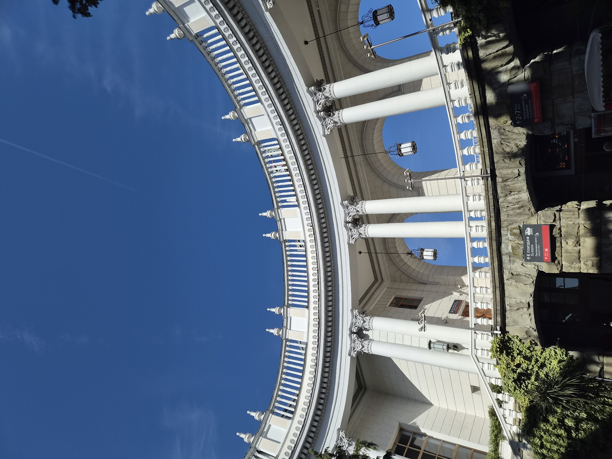 The image depicts a portion of the Sochi Railway Station in Sochi, Russia, with a view angled upwards towards the sky. A prominent feature is the semicircular colonnade, characterized by white columns topped with ornate capitals, supporting an archway. Decorative lamps hang beneath the arch. The sky is a clear, bright blue. To the right, the rough stone facade of the station's entrance is visible, adorned with signage, including "К поездам" and "To trains," indicating access to the trains. Some greenery is present along the base of the structure. The image showcases the station's architectural details and the surrounding environment.