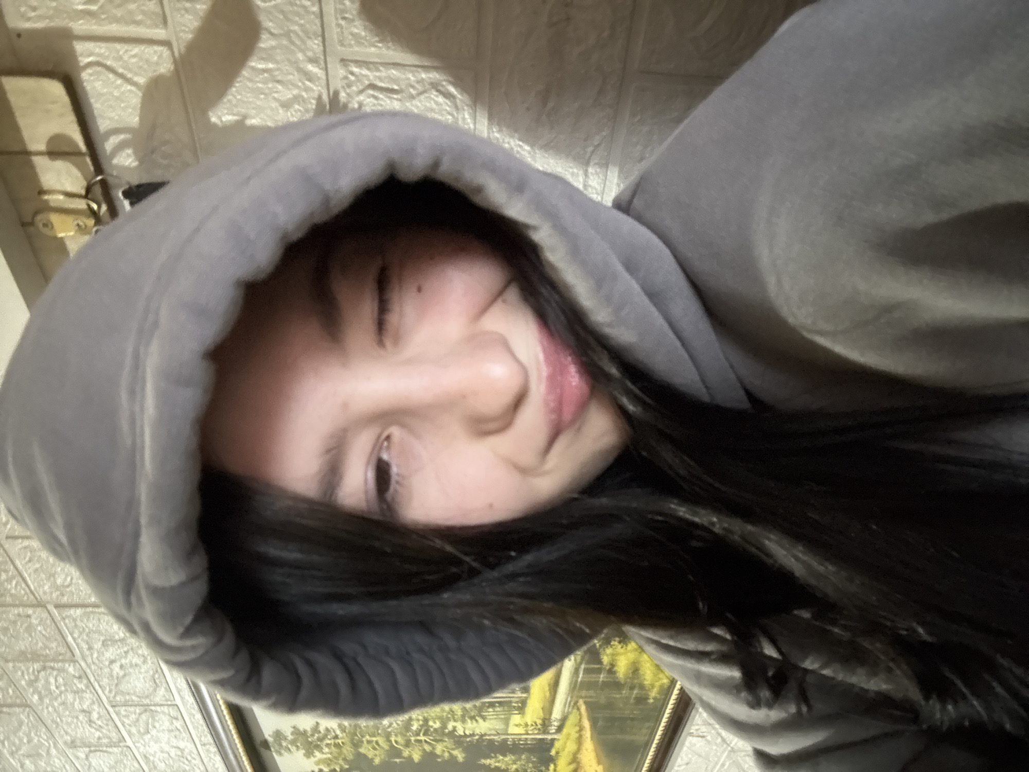 A young person with dark hair is wearing a gray hooded sweatshirt. The person's face is partially obscured by the hood. The person is looking directly at the camera. The background is a light-colored wall with a slightly textured pattern. A picture frame is visible at the bottom of the frame. The image is tilted. It is not possible to identify any location information in Фоки, Russia from the image.