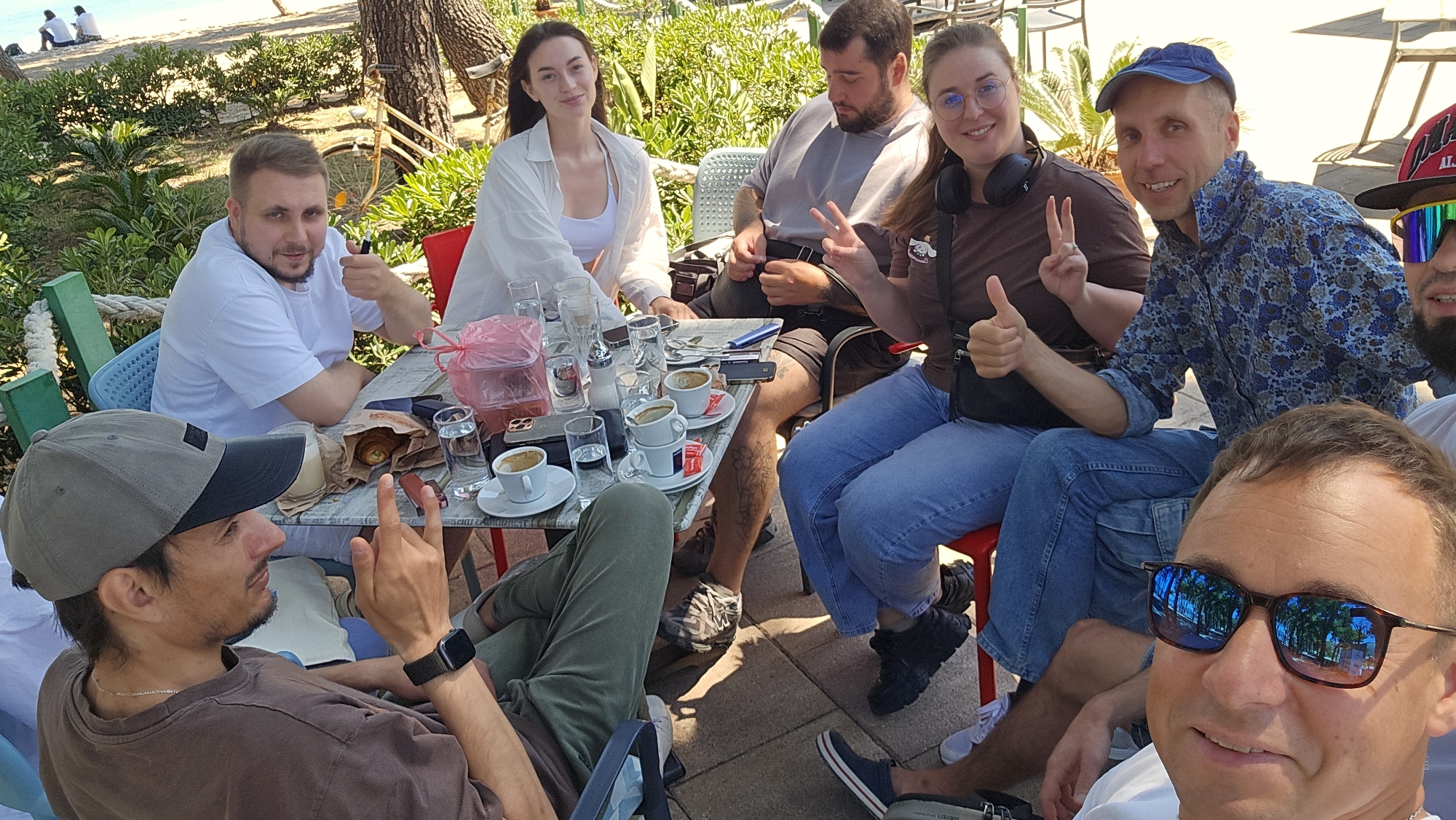 A group of eight people are sitting around a table outdoors, likely at a cafe, in Bar, Montenegro. The scene appears casual and relaxed, with most individuals smiling and posing for the camera. The table is covered with glasses, cups, and a plastic bag containing food. The foreground reveals a smiling man wearing sunglasses. To his left is a man in a grey cap pointing upward. Across from them is a man in a white shirt giving a thumbs-up. There is a woman in a white top and another person. There is a man wearing a brown shirt with headphones making the peace sign. Behind them is a man in a blue patterned shirt. To the left is a man with a beard wearing a cap. The environment consists of a beach in the background, shaded by trees, suggesting the cafe is near the coastline.