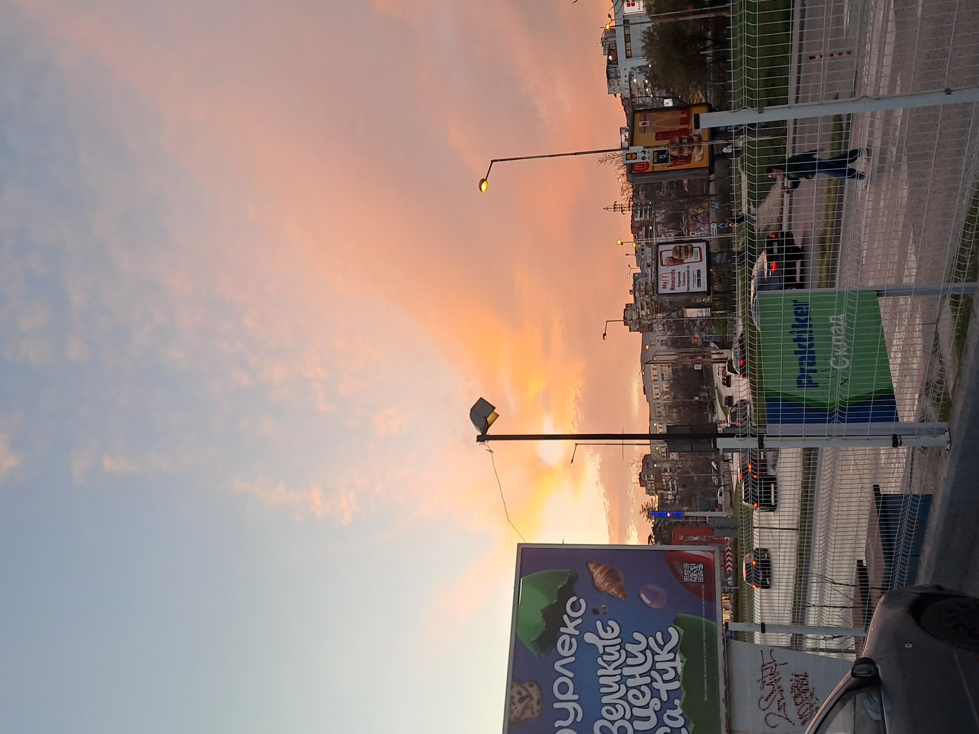 The image shows an urban scene in Veliko Tarnovo, Bulgaria, at dusk or dawn. The sky is a gradient of blue to orange-pink, indicating sunrise or sunset.
In the foreground is a billboard with Cyrillic script, advertising "Gourmet. Veliki ceni za tuk." A grey car is partially visible. A metal fence runs along the street. Behind the fence is a street with a pedestrian crossing. There are cars on the road, with their headlights visible.
There is a person crossing the street.
In the background, there are buildings, advertisements, and streetlights. The buildings are typical urban structures, and the advertisements are in Cyrillic, reinforcing the location in Bulgaria.