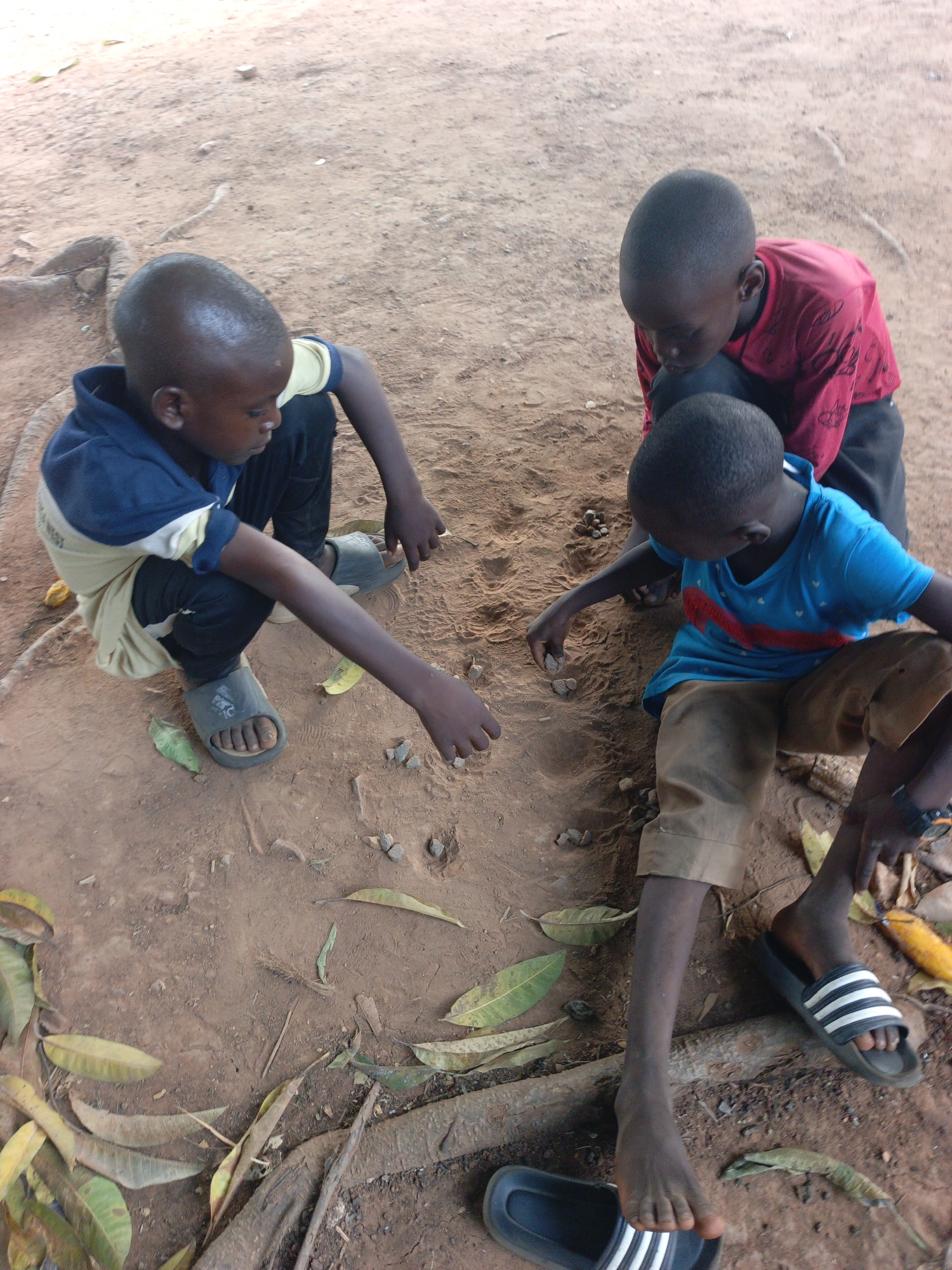 Three young boys are playing in the dirt in Akwanga, Nigeria. The ground is bare, reddish-brown earth littered with fallen leaves and small stones. One boy, wearing a blue and beige hooded shirt and black pants, squats on the left, reaching with his right hand towards the ground. He wears gray sandals. Another boy, wearing a blue shirt with a red stripe and tan pants, sits with his legs outstretched on the right. He is also reaching with his right hand. He wears black and white striped sandals. The third boy, wearing a red long-sleeved shirt and black pants, is leaning over the other two, looking down at the ground. A pair of black and white striped sandals is near the bottom right corner.