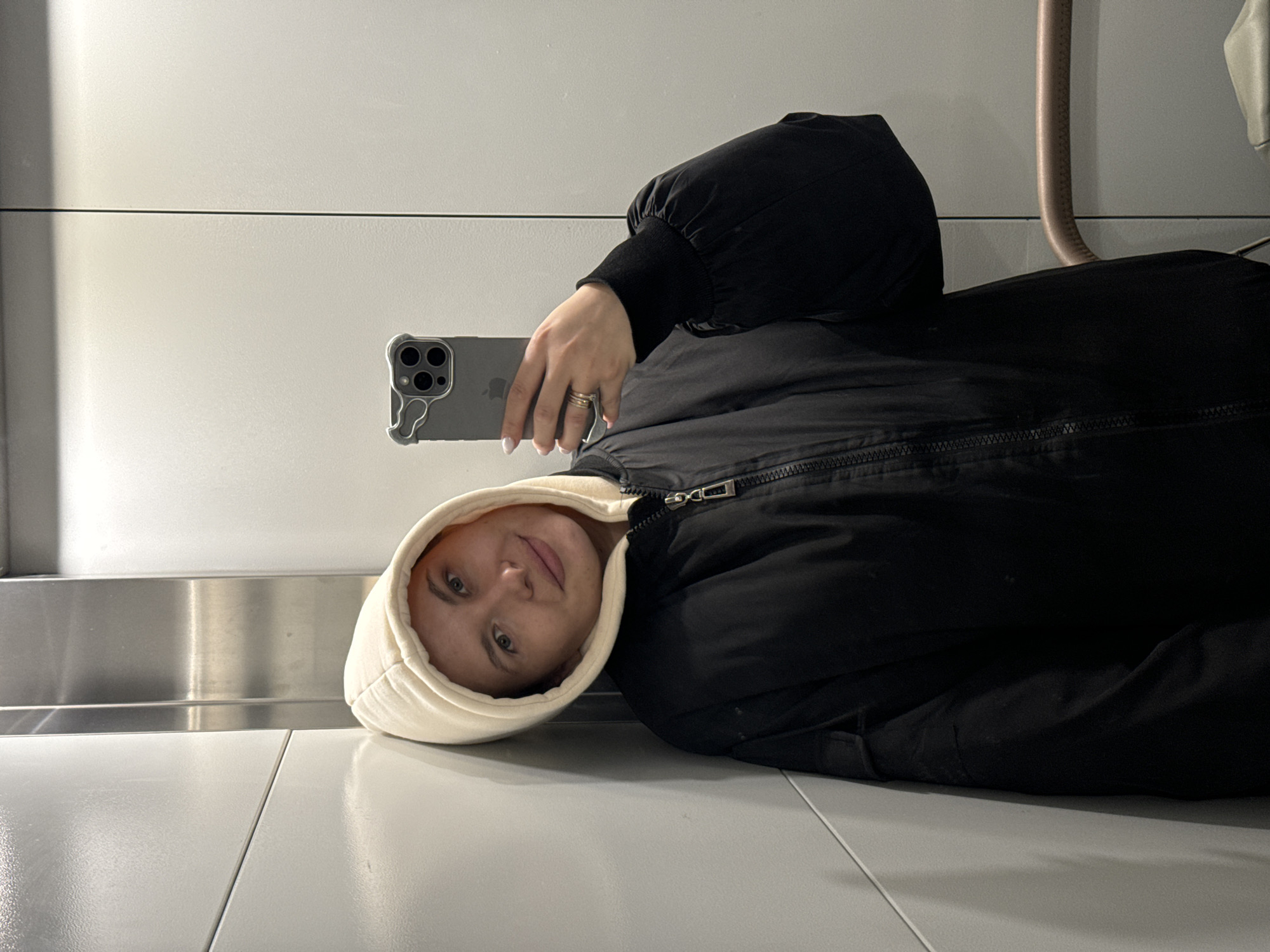 A person is depicted lying on their back, oriented perpendicular to the viewer, against a minimalist indoor background. The individual, who appears to be a young adult with light skin and light-colored eyes, is wearing a cream-colored hooded sweatshirt beneath a black puffer jacket with a visible zipper. They gaze directly at the camera with a neutral to slightly pleasant expression.
In their right hand, they hold a gray smartphone, which features multiple camera lenses on its back and a distinctive, wavy, possibly clear phone case. A ring is visible on their right ring finger. The Apple logo is faintly discernible on the back of the phone.
The setting consists of light gray or white tiled floors and matching walls, creating a clean, modern aesthetic. A dark horizontal line runs across the wall, suggesting a panel division or architectural feature. In the upper right background, a curved, light brown pipe or handle is visible. The lighting is bright and even, indicative of an indoor environment, possibly a public space like an airport or modern building interior. The person seems to be taking a selfie or recording themselves while lying down.