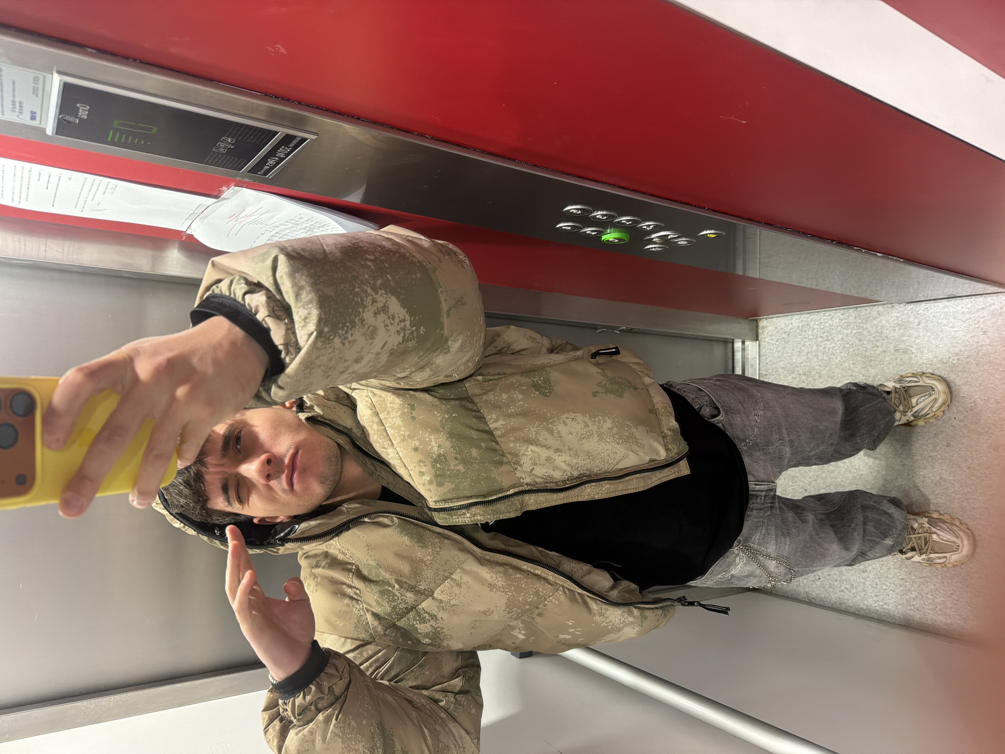 A young man is captured taking a selfie in an elevator, holding a yellow-cased smartphone in his right hand and looking directly into its camera. He wears a camouflage-patterned puffy jacket with a hood, a black undershirt, faded grey jeans, and light-colored sneakers. His dark hair is visible beneath the hood, and his expression is neutral.
The elevator interior features a vibrant red wall with a metallic strip running horizontally across it. Below this strip, a control panel is mounted, displaying various silver buttons, one of which glows green, indicating an active selection or function. A digital screen on the panel shows the word "OASIS" and below it, text that appears to include "230V / 60Hz" along with other symbols and numbers. Adjacent to the control panel, a document or sheet of paper with text is tucked into a slot. The opposite wall and the floor are a uniform light grey or silver, reflecting the indoor lighting. The overall scene suggests an ordinary moment inside an operational elevator, potentially in Sofia, Bulgaria, as indicated by the location context.