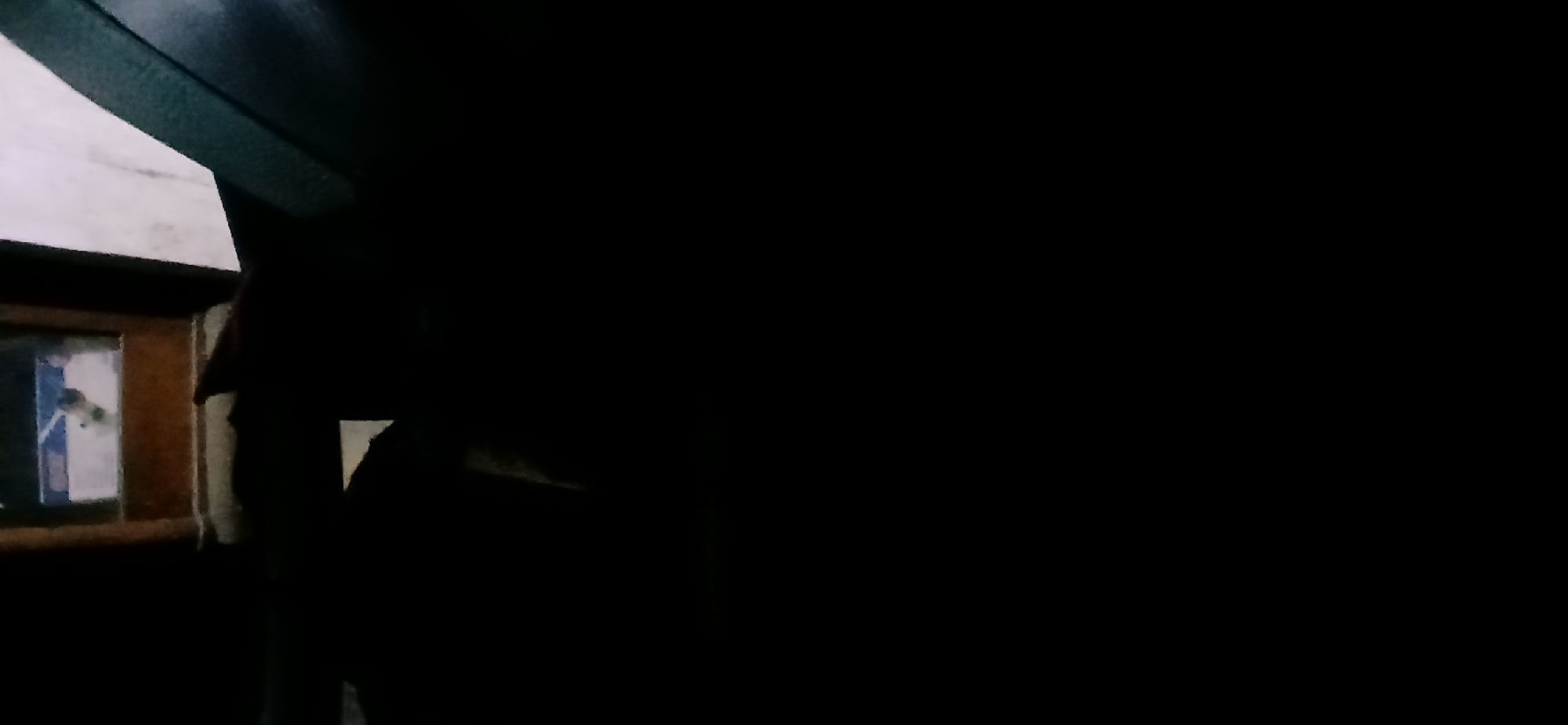 The image depicts a dark interior scene, possibly a room in a house or a vehicle. In the upper portion of the image, a dimly lit rectangular object with wooden framing is visible, which might be a window or a doorway. Inside this frame, a white object with some blue and dark markings is discernible, though its exact nature is unclear due to the low light conditions. To the right of this frame, a section of a dark, textured surface is present, possibly a wall or upholstery. The rest of the image is predominantly black, indicating a lack of significant light and rendering most details indiscernible. There are no clearly identifiable people or specific objects that would allow for precise scene localization within Kademangan Kulon, Indonesia. The overall impression is one of low visibility and a confined indoor space.