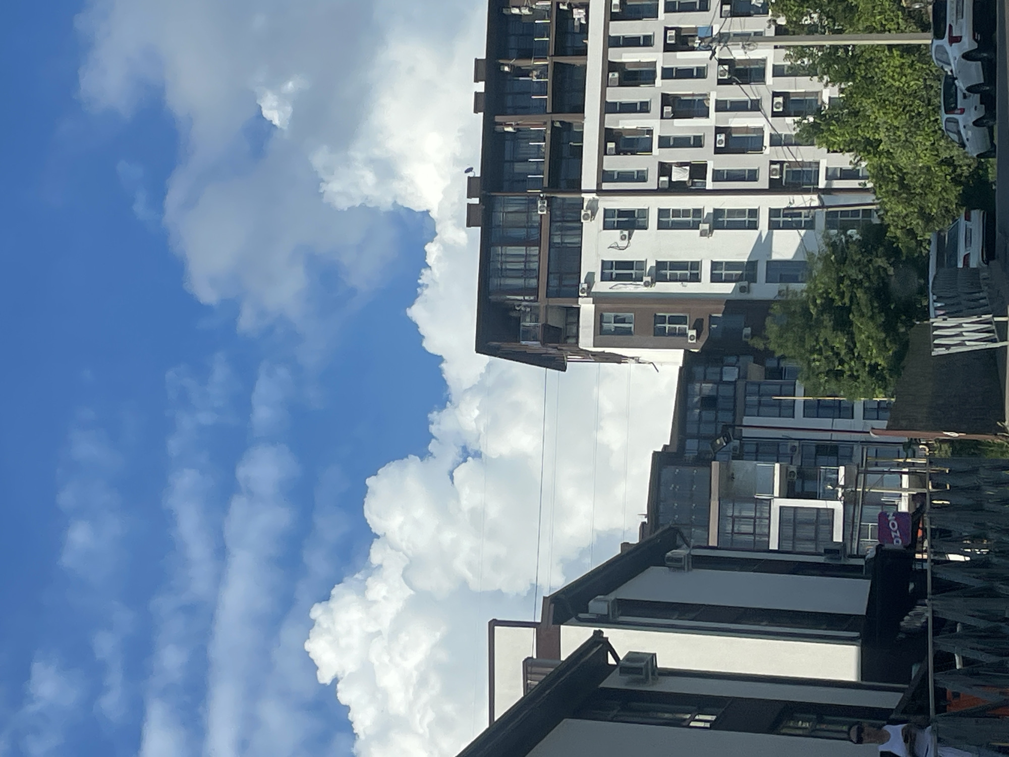 The image shows a street view in Sochi, Russia, with modern apartment buildings dominating the scene under a bright blue sky with fluffy white clouds. In the foreground, a dark fence partially obstructs the view of the street and sidewalk. To the left, a portion of a white building with dark trim is visible, featuring several air conditioning units. A woman in a white top and sunglasses stands near this building. Further down the street, a large, multi-story apartment building with a dark brown roofline and numerous glass-paneled balconies stands prominently. This building has a light-colored facade with dark vertical accents and many visible air conditioning units. To the right of this building, another apartment complex with a lighter facade and numerous windows is visible, with trees and vegetation in front of it. Several cars are parked along the street, including white sedans. A sign with the Cyrillic word "OZON" is visible on one of the buildings. The overall impression is a modern urban environment on a sunny day.