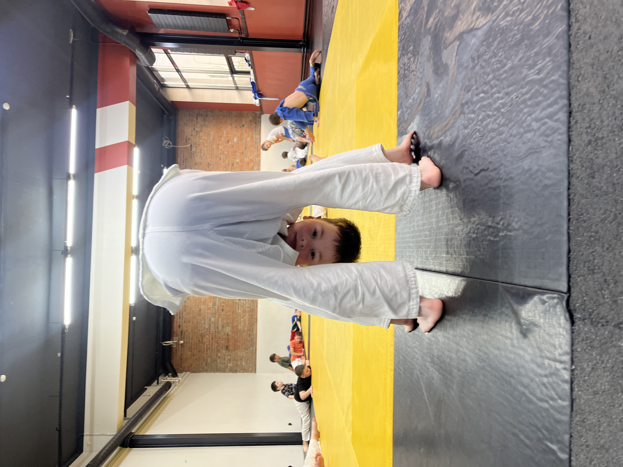 In a dojo with a yellow and grey mat, a group of children in white gis are practicing martial arts. The foreground is dominated by a child in a white gi bent over, facing the camera, with their feet visible. In the background, several other children are scattered across the mat. Some are in similar white gis, while others are in blue gis. The dojo features a red and white striped pillar, brick walls, and fluorescent lighting. The scene appears to be an indoor training session.