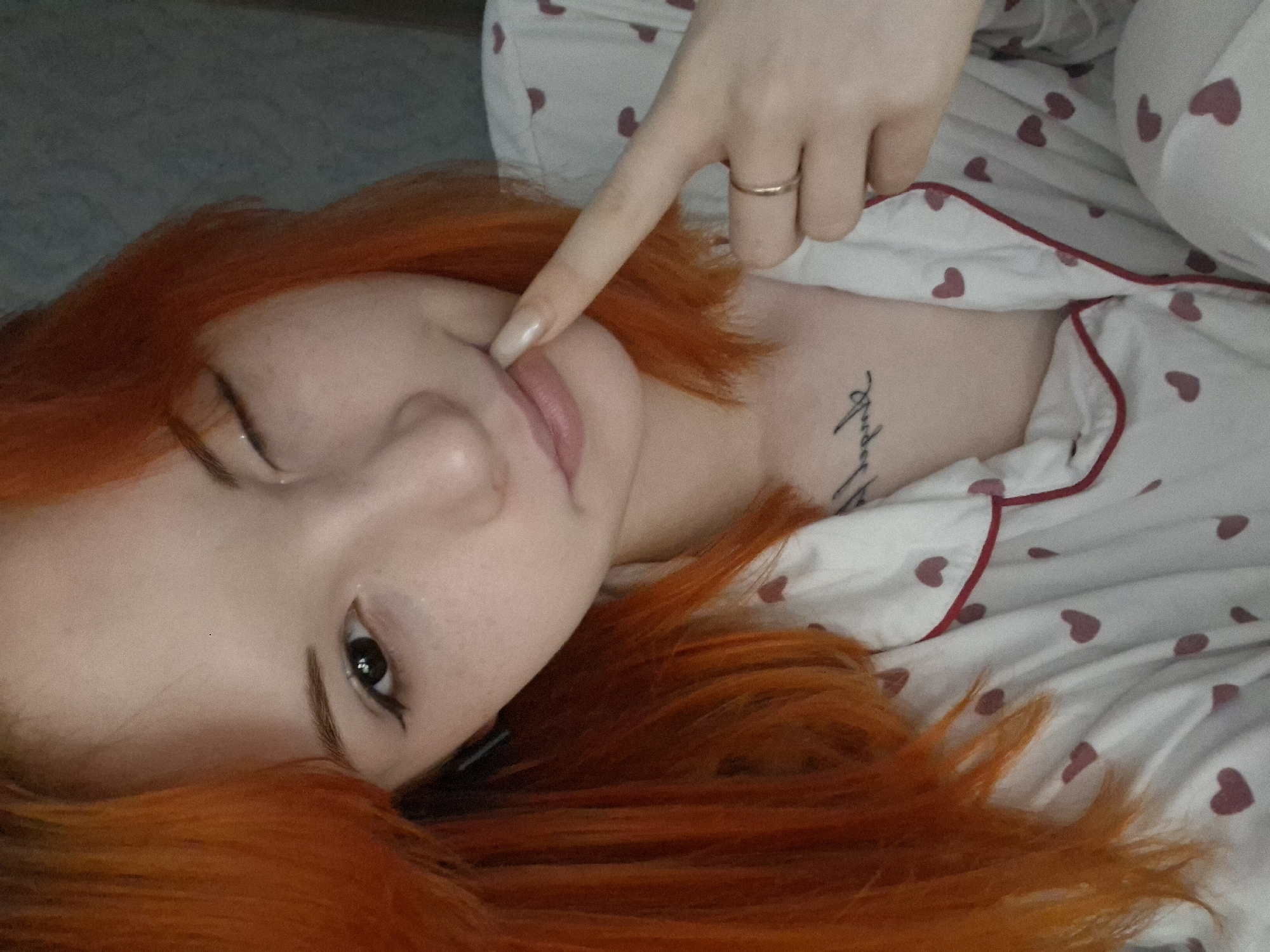 A pale-skinned woman with vibrant orange hair lies on a surface covered in white pajamas or bedding patterned with small, dark pink hearts trimmed with red. Her head is tilted to the side, positioning her face towards the camera, and her left eye is partially closed in what appears to be a wink. Her right eye is open, revealing dark lashes and liner. A hand with long, light-colored nails rests near her face, with one finger pointing towards her cheek. A simple gold band adorns one finger.
A tattoo of cursive writing is visible on her chest, just below the neckline of the pajamas. The background is blurred and indistinct, appearing as a gray surface or wall. There is no verifiable information about the location being in Chaikovsky, Russia.