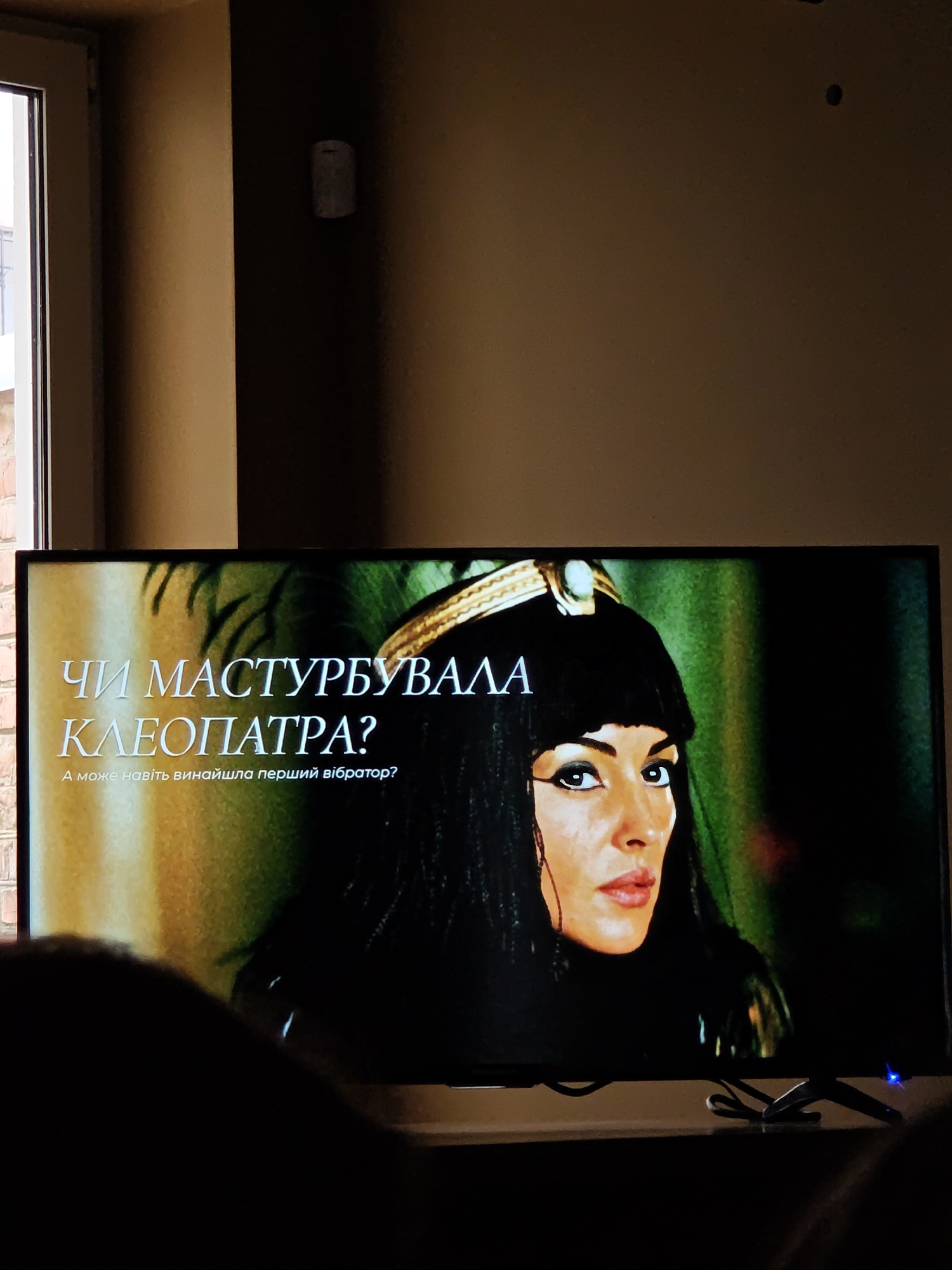 On a television screen, the question "Чи мастурбувала Клеопатра?" ("Did Cleopatra masturbate?") is displayed in Ukrainian. Beneath the question, it reads "А може навіть винайшла перший вібратор?" ("Maybe even invented the first vibrator?"). An image of a woman dressed as Cleopatra accompanies the text. The scene is captured indoors. The location, Vinnytsia, Ukraine, cannot be determined from the visual information provided.