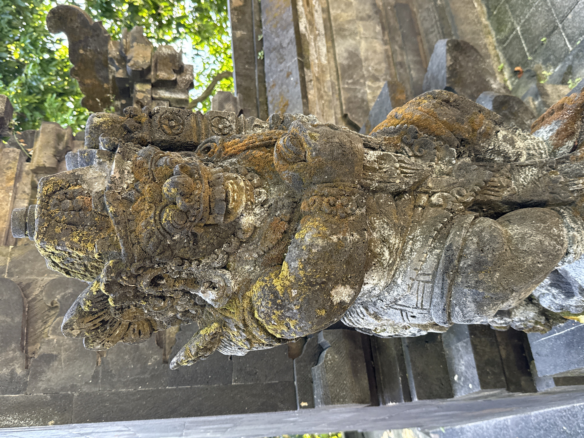 The image displays an ancient, intricately carved stone statue, likely depicting a guardian deity or mythical creature, characteristic of Balinese or Indonesian temple architecture. The statue is crafted from dark, weathered stone and is heavily adorned with extensive patches of green moss and yellowish-orange lichen, highlighting its age and prolonged exposure to the elements.
Notable features of the statue include an elaborate headpiece or crown, detailed facial features, and a robust body. One visible hand is clearly clenched into a fist, suggesting strength or a protective stance. The surface is covered with intricate carvings, featuring various patterns that could represent scales, decorative clothing, or other ornate motifs. On one side of the statue, there are etched lines or stylized symbols, appearing as geometric patterns.
The background reveals blurred elements of dark grey, rough-hewn stone architecture, such as pillars and walls, consistent with a temple structure. Lush green foliage and tree leaves are visible in the upper and far background, indicating an outdoor, natural setting. The scene is brightly illuminated by natural light, suggesting it is daytime with clear or sunny weather conditions. The setting is consistent with the provided location context of Kuta Selatan, Indonesia, showcasing a blend of ancient craftsmanship and tropical weathering.