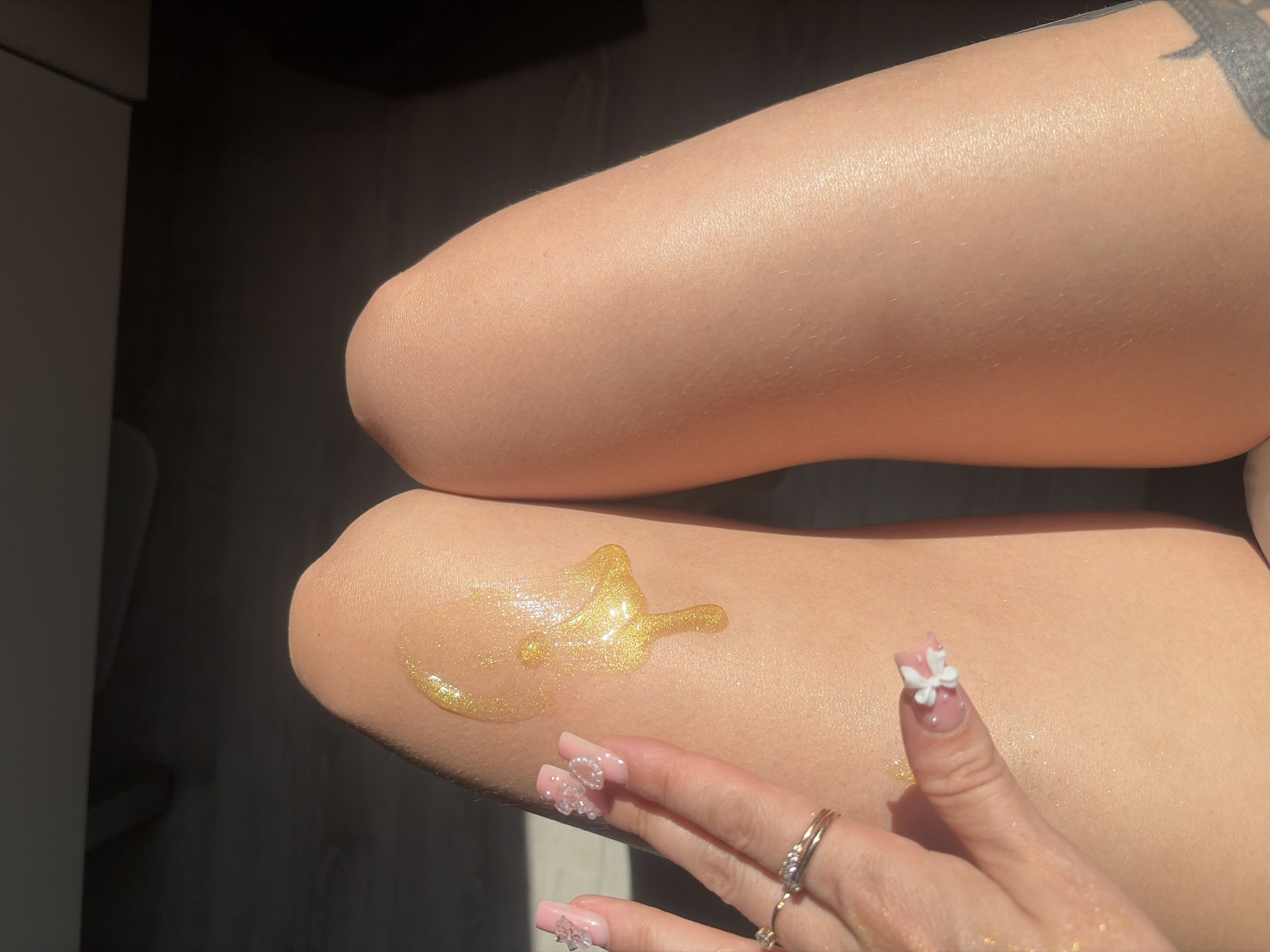 The image displays a close-up view of a person's legs, with a hand gently touching the skin. A viscous, golden liquid with shimmering particles is being applied or has just been applied to the left thigh, forming a droplet shape. The person's fingernails are decorated with pink polish and small white floral embellishments. A ring is visible on one of the fingers. The skin appears illuminated, likely due to the product. The background is out of focus, showing a dark, textured surface that could be a floor or furniture. Sunlight casts a shadow across the scene, suggesting an indoor setting with natural light. There are no discernible landmarks or clues indicating a location in Saint Petersburg, Russia.
