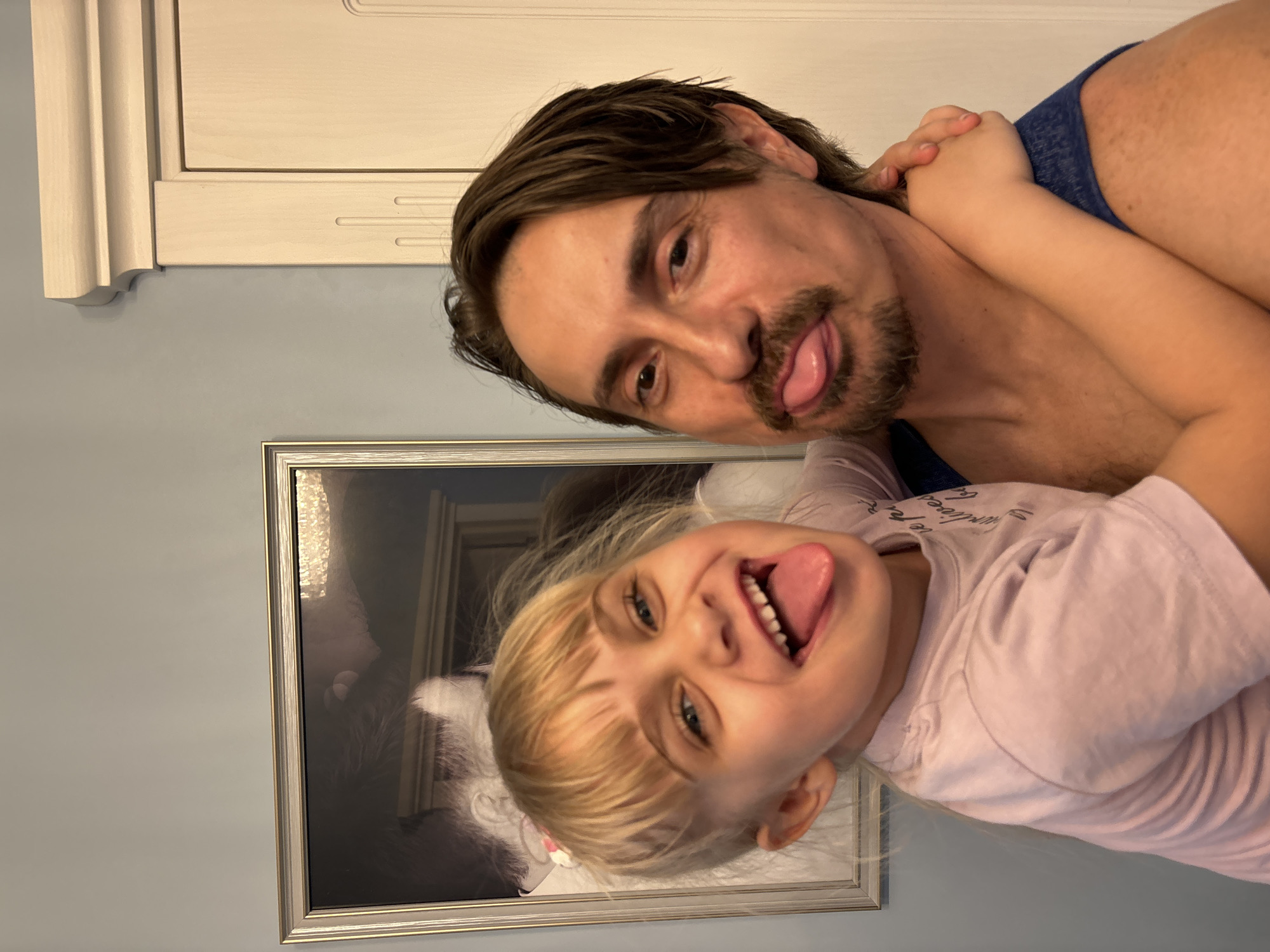 The image shows a father and his young daughter making silly faces for the camera. The father, with dark hair and a mustache, is leaning in, sticking his tongue out. The daughter, with blonde hair, is also sticking her tongue out and smiling. They appear to be in a home setting, possibly a living room or bedroom, with a framed picture on the wall behind them. The picture on the wall appears to be a portrait of a cat. The lighting suggests it is daytime. The mood is playful and affectionate. No visible text is present in the image.