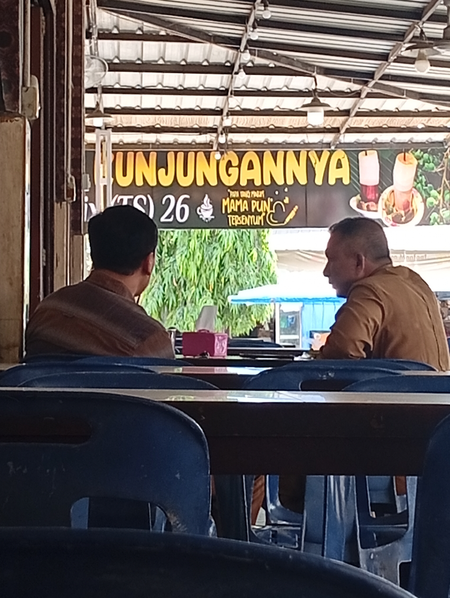 The image depicts an outdoor seating area at a food establishment in Lhokseumawe, Indonesia. Two men are seated at a table, their backs to the camera. The man on the left wears a brown, striped shirt, and the man on the right wears a brown uniform. In front of them, on the table, are a pink tissue box and a small pink object, possibly a lighter or a small container. Behind them, a large banner with yellow text reads "UNJUNGANNYA" and some Indonesian text below. Two drinks, one red and one pink, are displayed on the banner. The environment appears to be a covered outdoor space with a corrugated metal roof supported by wooden beams. Green foliage is visible in the background, suggesting a tropical climate. The seating area is furnished with blue plastic chairs.