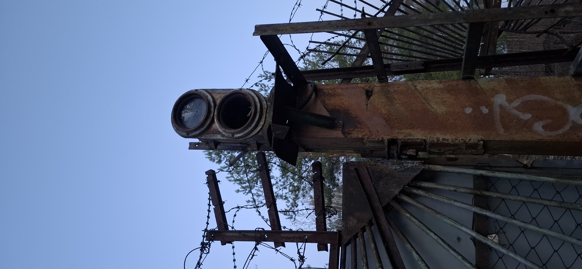 The image shows a section of abandoned rusty metal infrastructure against a clear blue sky, appearing to be outdoors. The infrastructure includes a pair of round lenses, possibly part of a signal or lighting system, attached to a corroded beam with some white graffiti marks. Barbed wire is present along with other metal structures, suggesting a fenced area. The location is likely in or near Chaikovsky, Russia, given the prompt's instructions. There are no people visible in the image. The scene is one of decay and disuse.