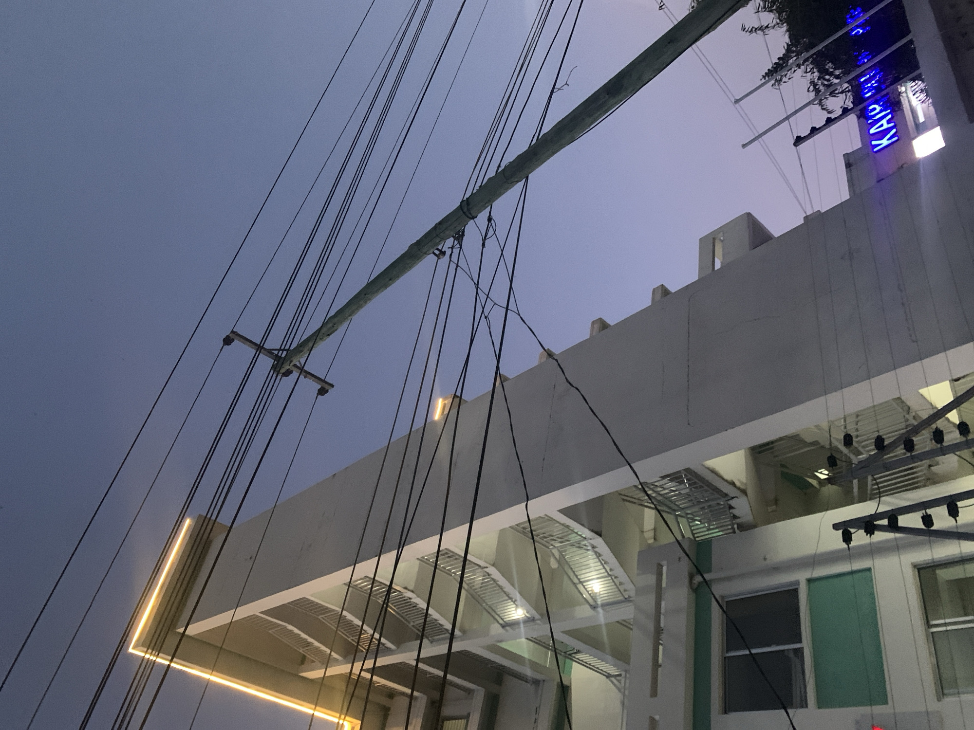 The photograph shows a low-angle view of a building exterior with a grey sky in the background. A weathered utility pole cuts diagonally across the frame, with numerous black wires extending from it, obscuring parts of the structure. The building is predominantly white, featuring a modern design with multiple tiers and projecting sections. Linear, recessed lighting illuminates the underside of some of the tiered sections, creating a warm, yellow glow. A neon sign, partly visible on the upper right of the building, displays blue lettering. The building's exterior also includes white window frames and green walls in the lower right area. There are no people visible in the scene. The building and wires suggest an urban setting, potentially in Dar es-Salaam, Tanzania.