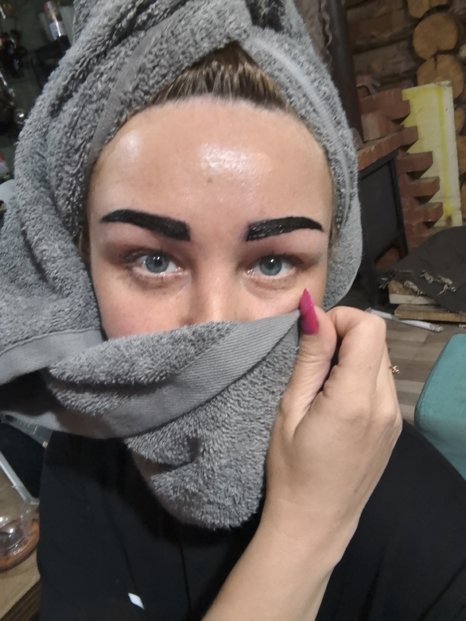 A selfie shows a person with light blue eyes and a gray towel wrapped around their head, also covering the lower half of their face. Their eyebrows are thick and dark. The person’s left hand, featuring a bright pink fingernail, is holding the towel. A portion of a woodpile can be seen in the background. The location cannot be verified.