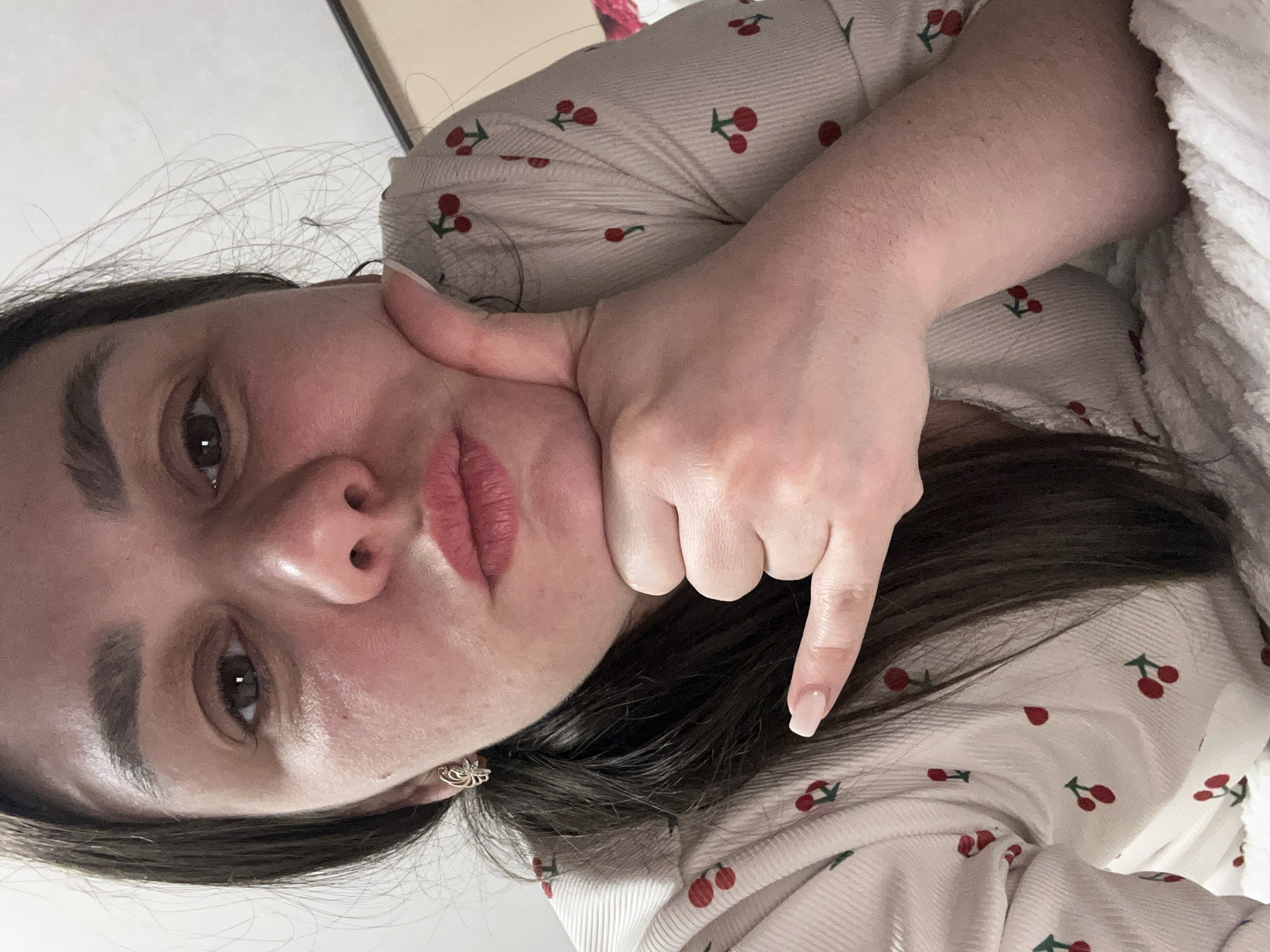 A woman with dark hair and brown eyes is shown in a close-up, angled view. She's wearing a light-colored shirt with a cherry pattern. Her left hand is touching her cheek, while her right hand is positioned with the index finger pointing downwards. She is wearing a silver-colored floral earring. The background is blurred. The location is unknown and there is no evidence it is located in the city of Chaikovsky, Russia.