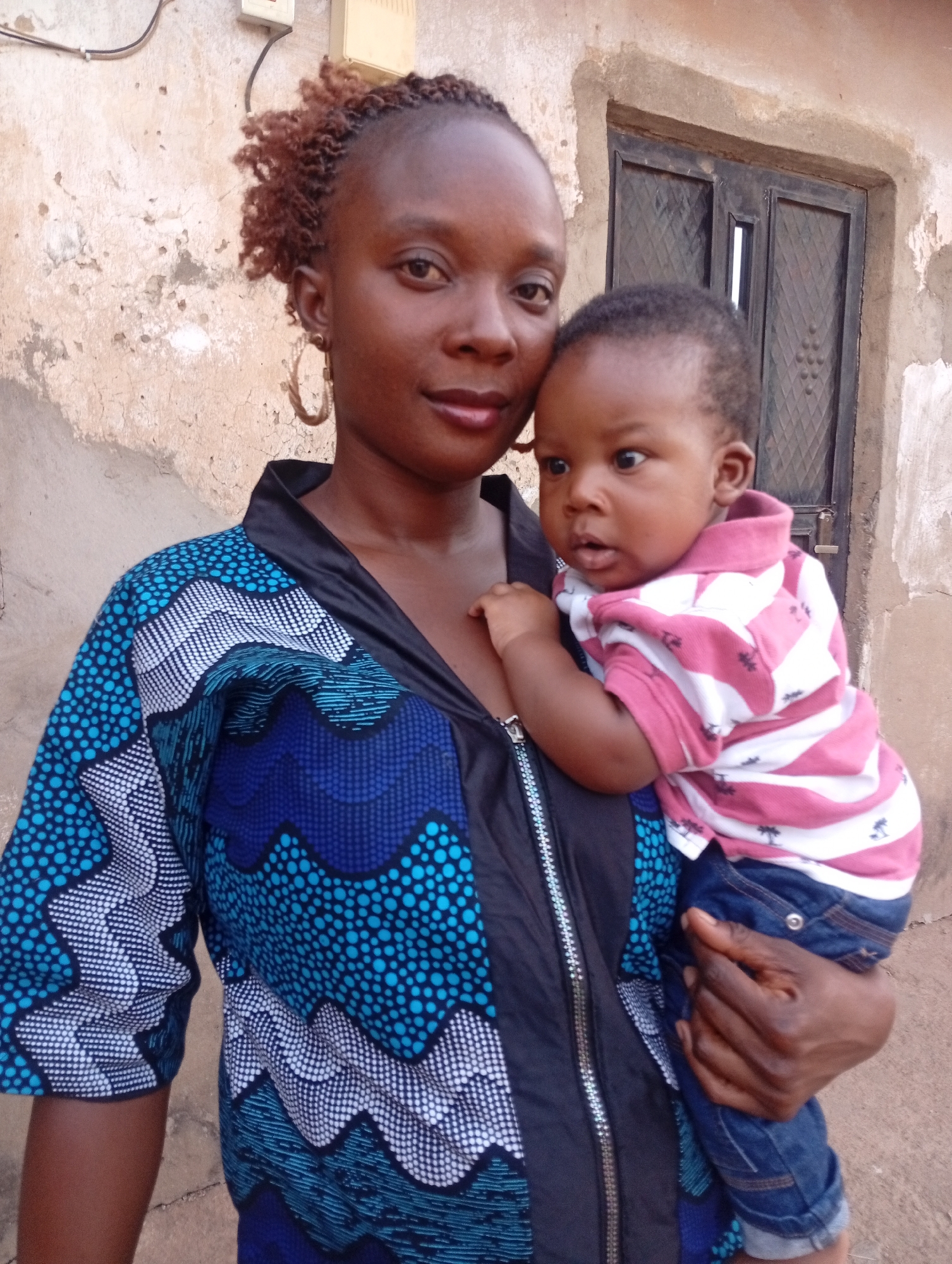 A woman holds a baby in front of a textured wall. The woman is wearing a blue and white patterned jacket over a black top. She has dark skin, and her hair is styled in reddish-brown braids. She is wearing large hoop earrings. The baby is wearing a pink and white striped shirt and blue jeans. The background shows a textured wall, and a black window frame in the upper right. Sabon Tasha, Nigeria can be inferred from the ethnic appearance of the people in the image and the style of the clothing.
