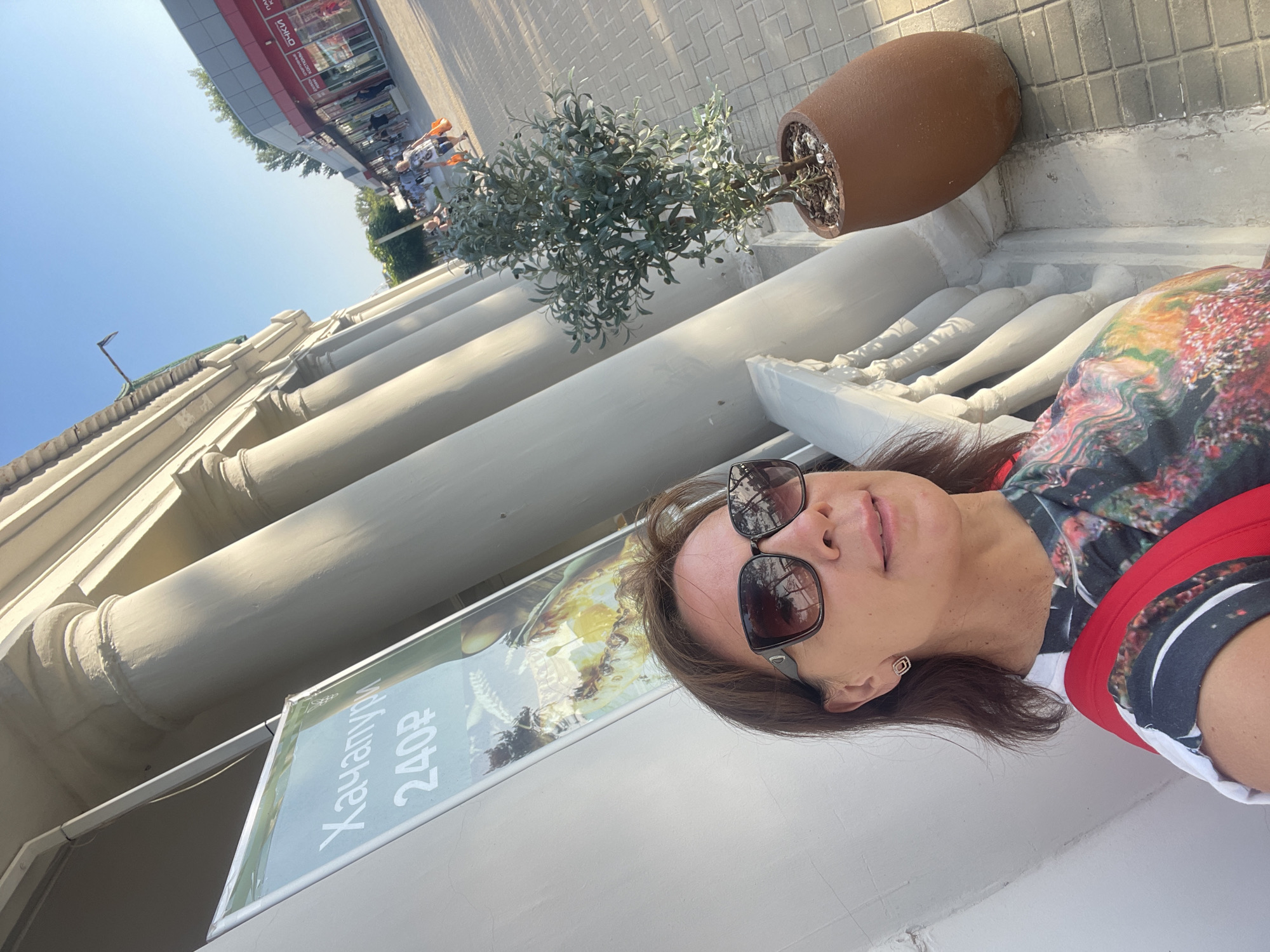 This media file is a first-person, low-angle, outdoor photo taken on a sunny day. The subject is a woman with brown hair wearing sunglasses and a colorful, abstract-patterned shirt with a red strap visible. She appears to be looking up and smiling slightly. In the background, there is a white balustrade and a large brown planter with a small tree or bush. Further back, a building with arched windows is visible, and beyond that, a row of shops with signage. A street or sidewalk area with tiled paving is also present. A poster or sign advertising "Хачапури 240₽" (Khachapuri 240 rubles) is partially visible in the lower left portion of the frame, suggesting a food-related establishment. The scene appears to be a public walkway or promenade. The lighting suggests it is daytime, likely late morning or afternoon, given the bright sun and clear blue sky. The overall mood is relaxed and pleasant. The location is identified as Anapa, Russia.