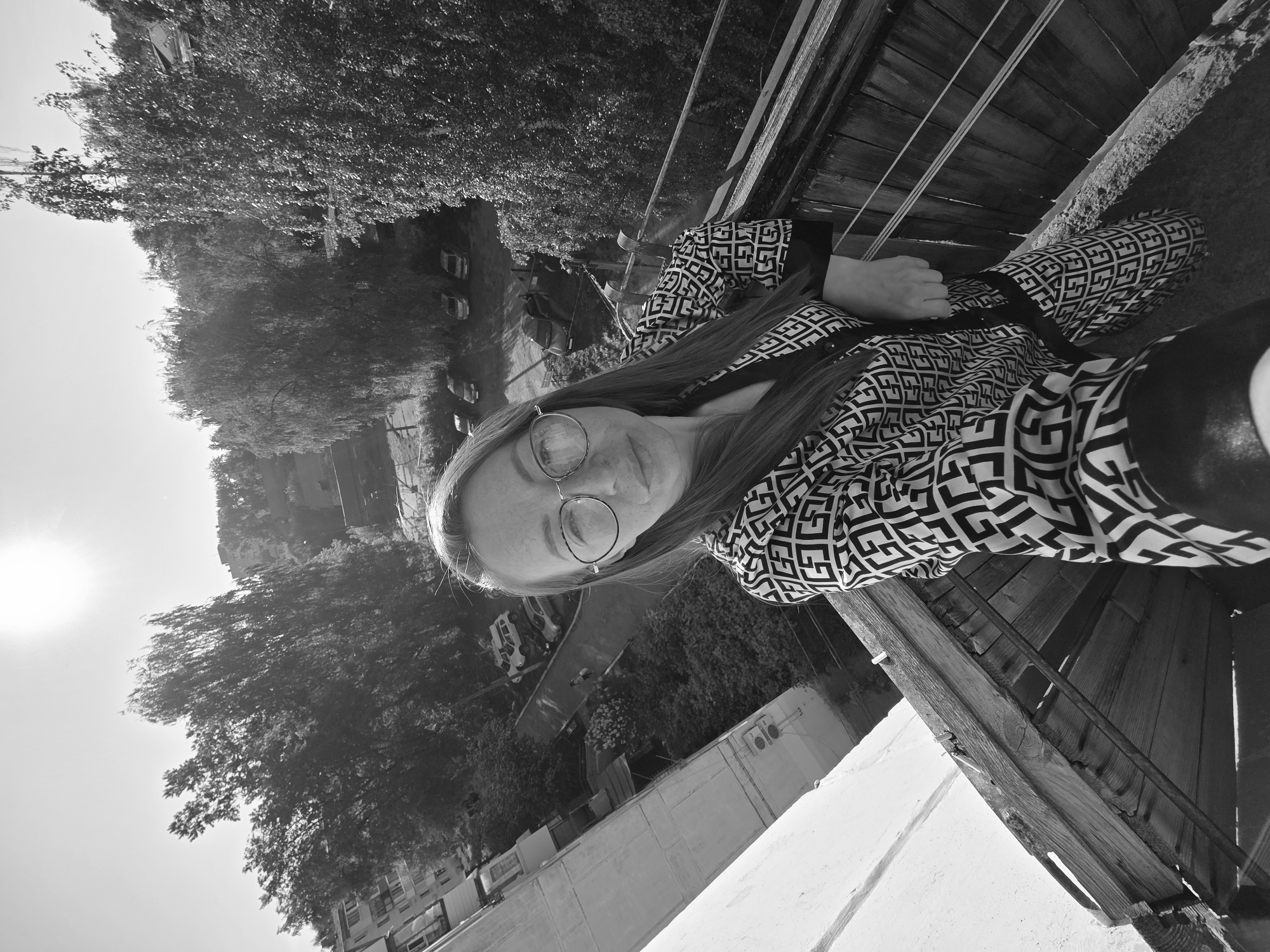 A black and white photograph depicts a young woman with long hair and glasses, wearing a patterned top. She is positioned on what appears to be a balcony with a wooden railing. In the background, trees and buildings are visible. Based on the available information, the location is Chaikovsky, Russia. The photo seems to be taken at ground level and oriented for social media posting.