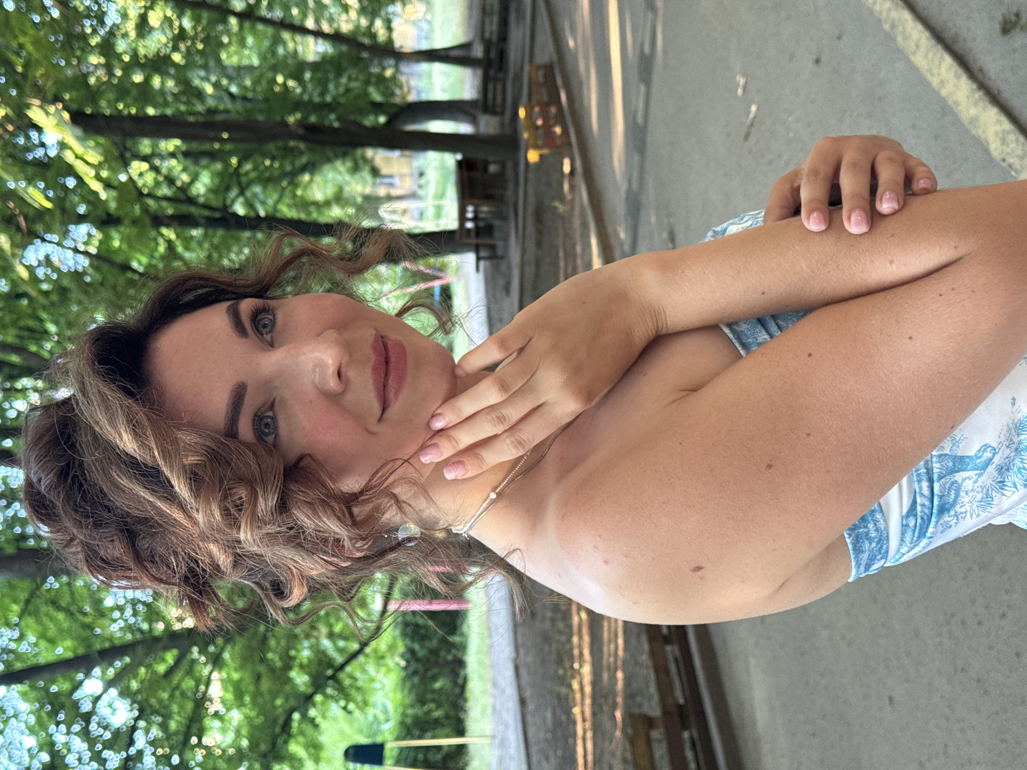 A woman is pictured from the chest up, with her face turned towards the camera. She has wavy brown hair and is wearing light makeup. Her left arm is bent, with her hand resting on her neck. Her right arm is crossed over her chest. She is wearing a blue and white patterned dress. The background appears to be an outdoor setting with trees and a concrete path. The lighting suggests it is daytime, possibly late afternoon due to the shadows. The image is taken in Kryvyi Rih, Ukraine.