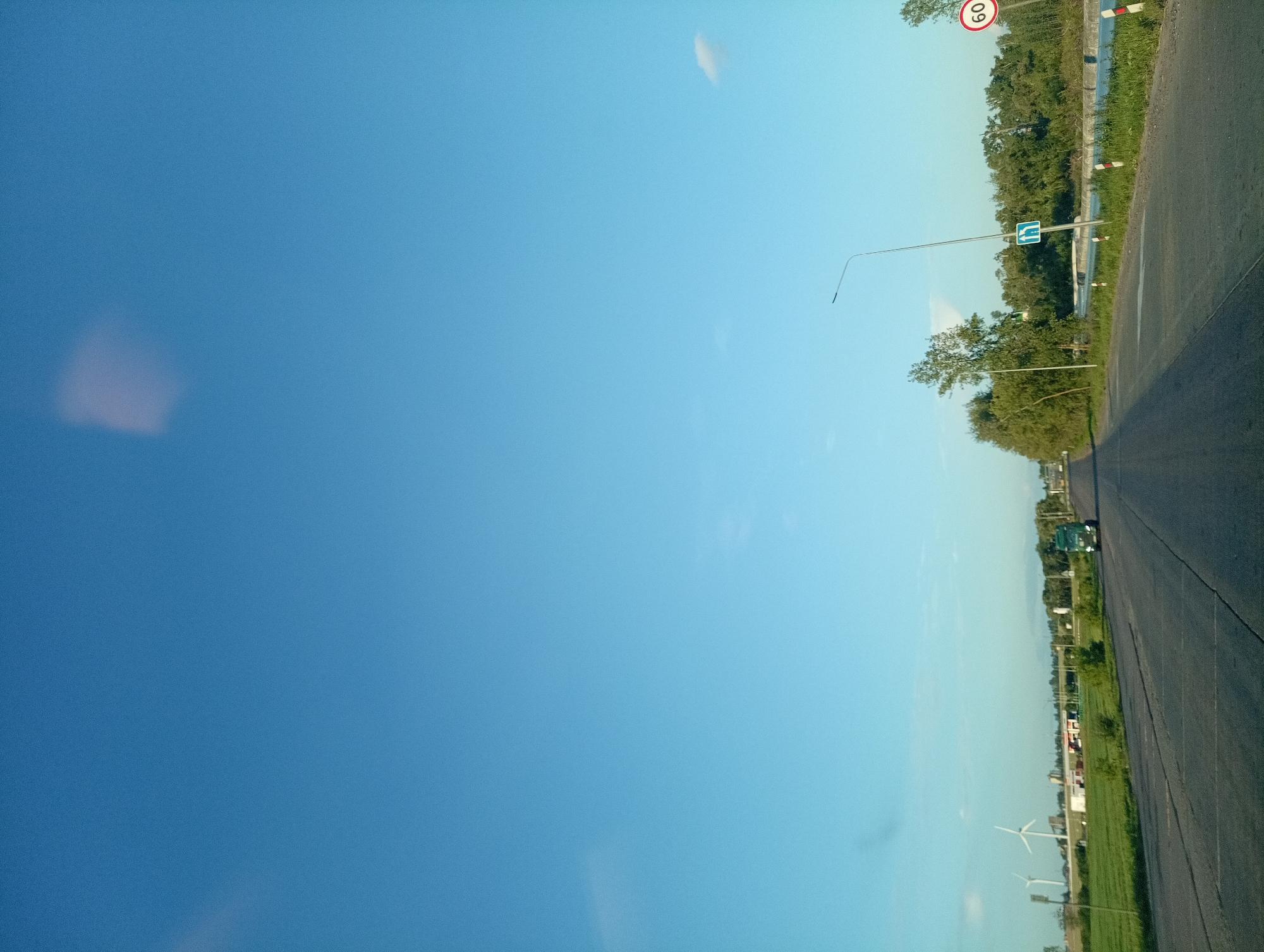 The image depicts a road scene in the vicinity of Вяйнянскі сельскі Савет, Belarus. The perspective is from a vehicle moving along a paved road. On the left side of the road, there is a grassy field with several wind turbines visible in the distance, along with some low-rise buildings. A green truck is visible on the road ahead, traveling in the same direction as the viewer. On the right side of the road, there is a body of water bordered by a metal guardrail, and a dense line of trees. A street light and a blue road sign indicating a directional change are also present on the right side of the road. A circular speed limit sign with the number "60" is also visible on the right. The sky is clear blue with a few faint clouds. The overall impression is of a rural or semi-rural landscape during daylight hours.
