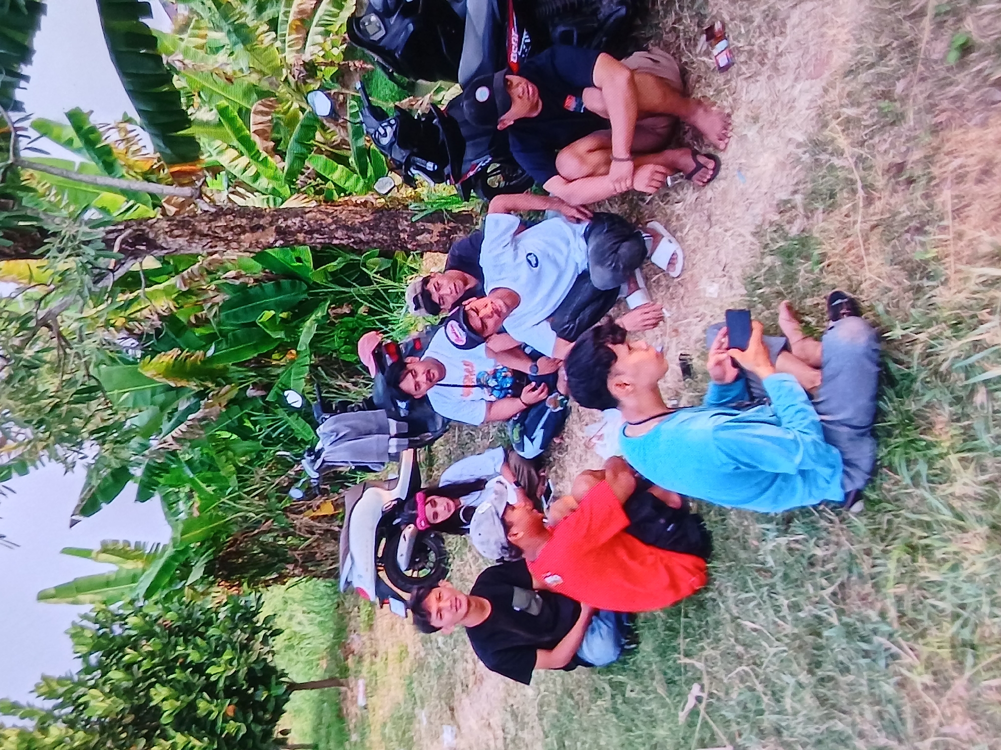In Setia Bakti, Indonesia, a group of eight young men are gathered outdoors, seated on the ground amidst lush vegetation and two parked motorcycles. The group includes individuals wearing t-shirts and various headwear such as baseball caps and a pink bandana. Some are looking at the camera, while others are engaged in conversation or looking at a mobile phone. The environment suggests a rural or semi-rural setting, with banana trees and other foliage forming a dense backdrop. The lighting indicates it is daytime, possibly late afternoon given the soft light. One of the motorcycles is white and appears to be a scooter, while the other is black. The ground is a mix of dirt and dry grass.