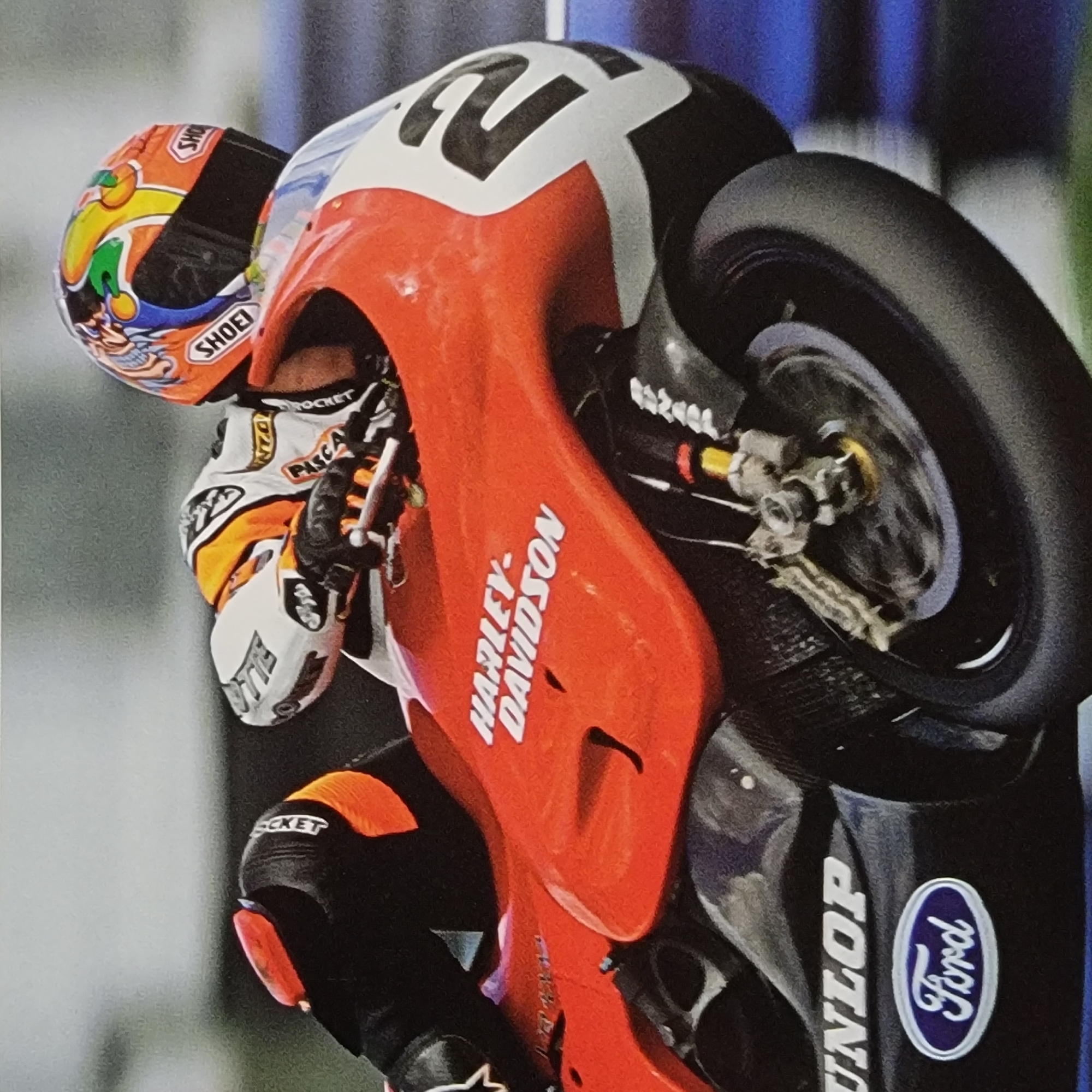 The image is a close-up, vertically oriented, low-angle shot of a motorcycle racer performing a wheelie. The racer is wearing a colorful, orange and yellow helmet with "SHOEI" visible. Their racing suit is primarily orange and white with black accents and features logos for "ROCKET" and "PASCAL". The motorcycle is predominantly orange with a large "2" on the fairing in white and black. "HARLEY-DAVIDSON" is prominently displayed in white lettering on the side of the motorcycle. The rear wheel and part of the braking system are visible. The motorcycle also displays a "DUNLOP" logo in white on a black surface, and a blue and white "Ford" oval logo. The background is blurred, with hints of blue and grey. The lighting suggests an outdoor setting, possibly a race track, with bright illumination. The focus is on the dynamic action of the wheelie and the branding of the racer and their equipment.