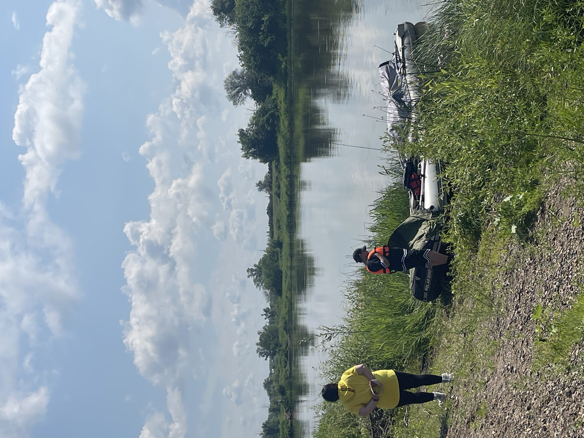 The image depicts a serene river scene with two individuals standing on the grassy bank. In the foreground, a person wearing a bright yellow hoodie and black leggings stands with their hands clasped behind their back, facing away from the camera. To their right, another individual in a black t-shirt with white stripes and an orange life vest is positioned near an inflatable boat. The boat, a "Polar Bird," is partially covered with a grey tarp and rests by the water's edge. A second, smaller inflatable boat is also visible. The river is calm, reflecting the lush green trees and a partly cloudy sky. The background shows a dense line of trees and vegetation along the opposite bank, with more trees and a field visible in the distance. The overall impression is of a tranquil, outdoor setting, likely a recreational area near Nezhinka, Russia.