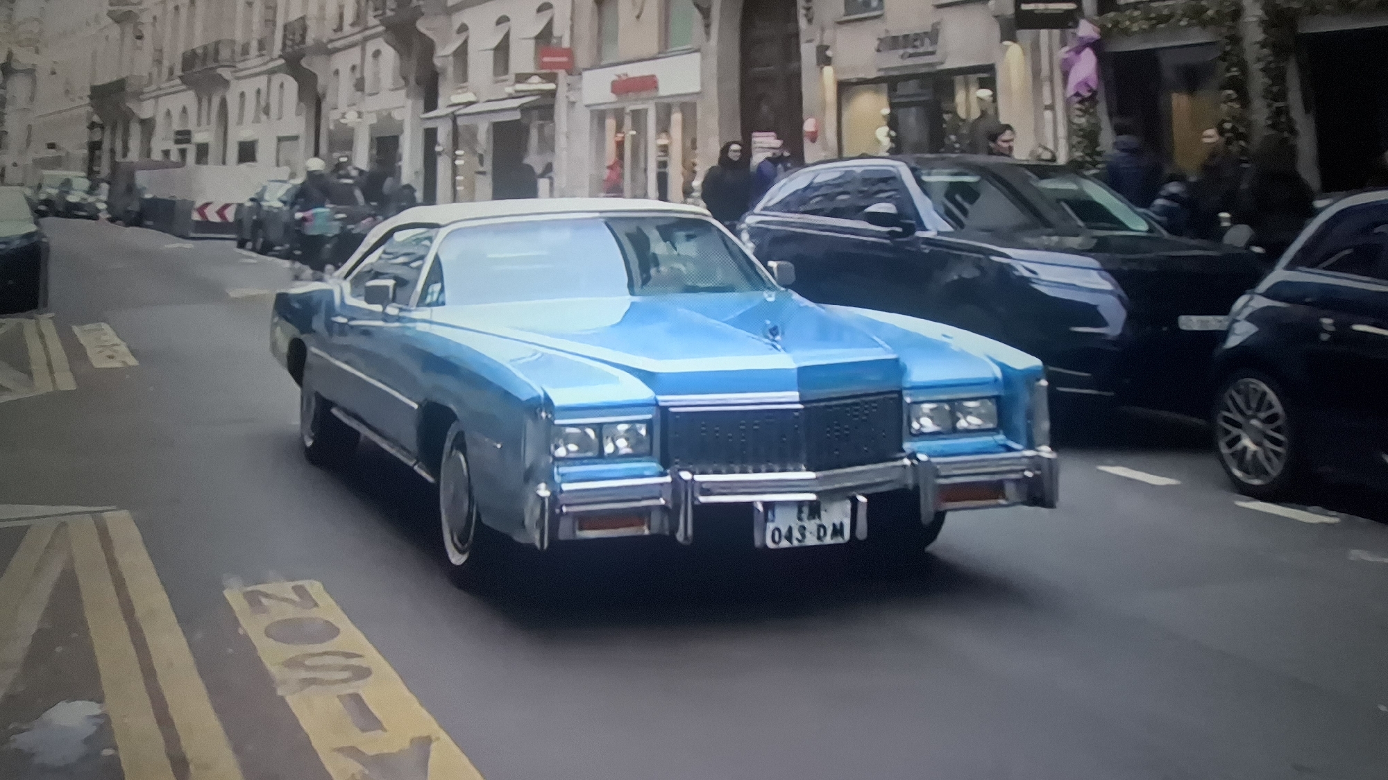 The image shows a light blue vintage car with a white top moving on a street in an urban area. The license plate reads "EW-043-DM." The street has marked parking zones. Behind the car, there are other vehicles and pedestrians on the sidewalks in front of shops and buildings. The city is currently unidentified.