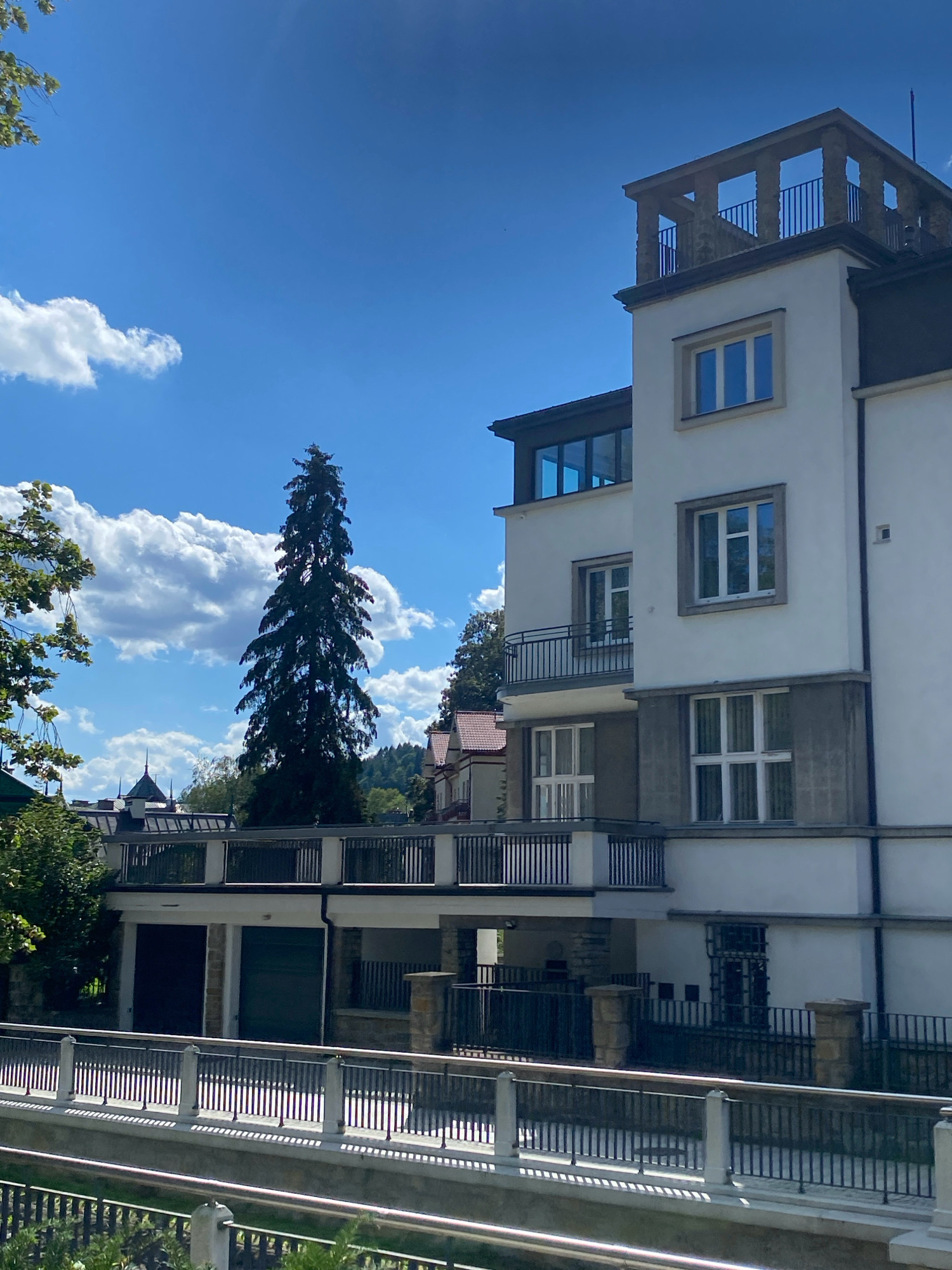 The image depicts a modern white building with multiple floors and balconies, situated in Krynica-Zdrój, Poland. The building features large windows, some with reflective blue glass, and a distinctive rooftop observation deck with stone pillars and railings. In the foreground, a black metal fence runs parallel to a walkway or a bridge, separating it from a body of water below, which is bordered by lush greenery. To the left of the building, a tall evergreen tree stands out against a bright blue sky dotted with fluffy white clouds. In the background, more buildings with red-tiled roofs and a distant hillside covered in trees can be seen. The scene is bathed in natural daylight, suggesting it was taken on a clear day. There are no people visible in the image.