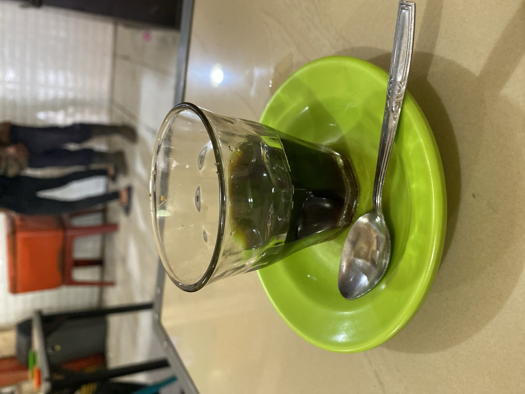 In Lhokseumawe, Indonesia, a close-up shot focuses on a clear glass filled with dark liquid, likely coffee, positioned on a bright green saucer. A silver spoon rests on the saucer, parallel to the edge of the frame. The setting appears to be an indoor establishment, possibly a cafe or eatery, with a light-colored tabletop beneath the glass and saucer. In the blurred background, there are hints of people and furniture, including a red cooler and white tiled walls, suggesting a public space.