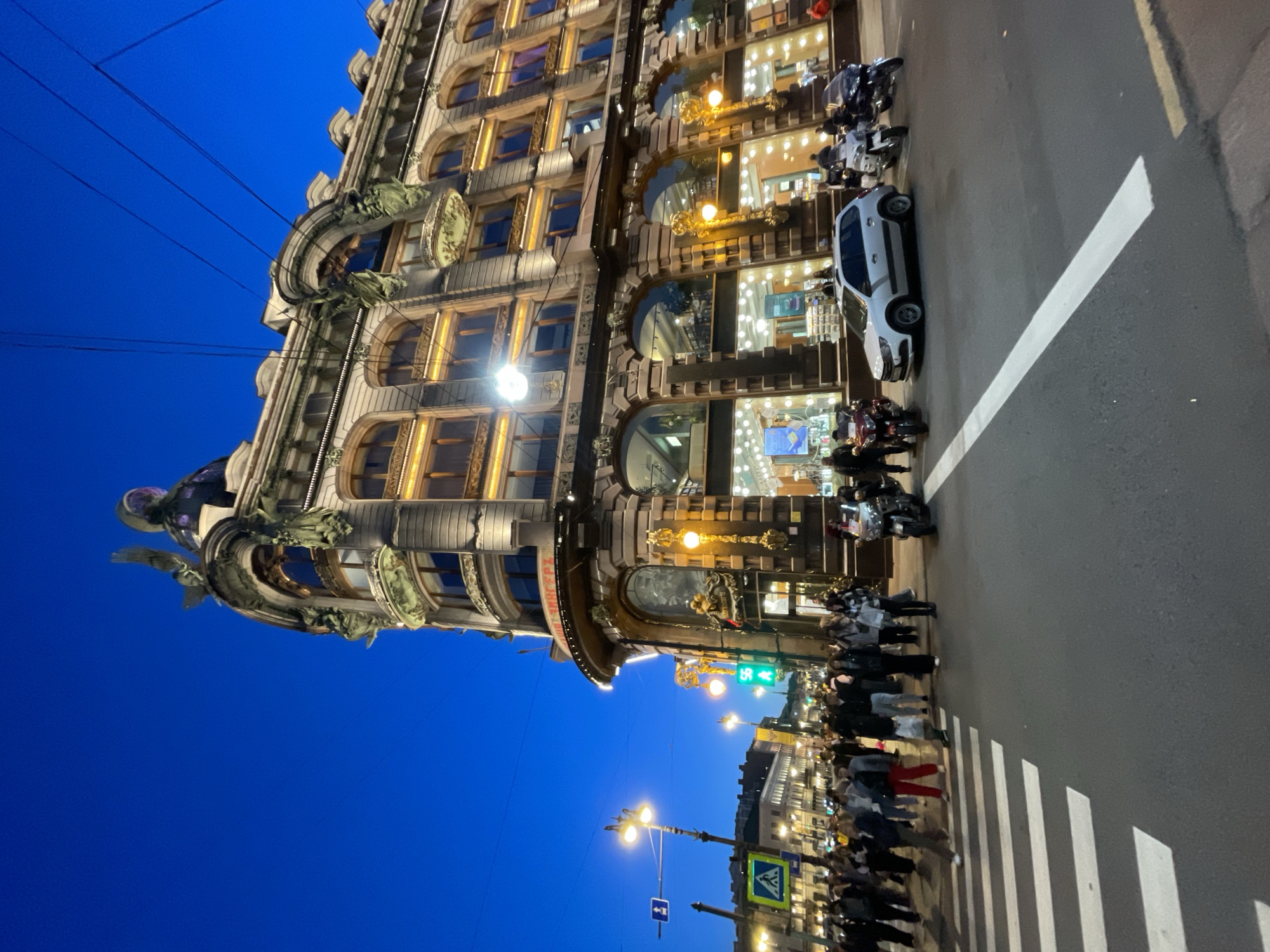 The image captures a vibrant urban scene at dusk or night, dominated by a magnificent, ornate building on the left side, illuminated by its interior lights and street lamps. The sky above is a deep, clear blue, indicating the transition from twilight to night.
The central subject is a grand, multi-story building featuring intricate Art Nouveau architecture, with a distinctive rounded corner. Its facade is richly detailed with arched windows, decorative cornices, and elaborate sculptures, including what appear to be mythical figures or gargoyles adorning its upper levels. The numerous windows emit a warm, golden glow, suggesting active businesses or displays inside. Overhead wires are visible against the deep blue sky.
At street level, a bustling crowd of people walks and gathers on the sidewalk alongside the building. A white car is parked on the paved street, along with several motorcycles. The street features a clearly marked zebra crossing with broad white stripes. Further down, more illuminated buildings line the street, accompanied by several streetlights. A green traffic light is visible in the distance, alongside a blue square directional sign with a white arrow and a triangular pedestrian crossing sign mounted on a pole.
A notable detail is the Cyrillic text visible on a curved section of the building's facade, which reads "ДОМ ЗИНГЕРА" (Dom Zingera), identifying it as the historic Singer House (or House of Books) in St. Petersburg, Russia. The scene conveys a lively, active city environment during the evening hours.