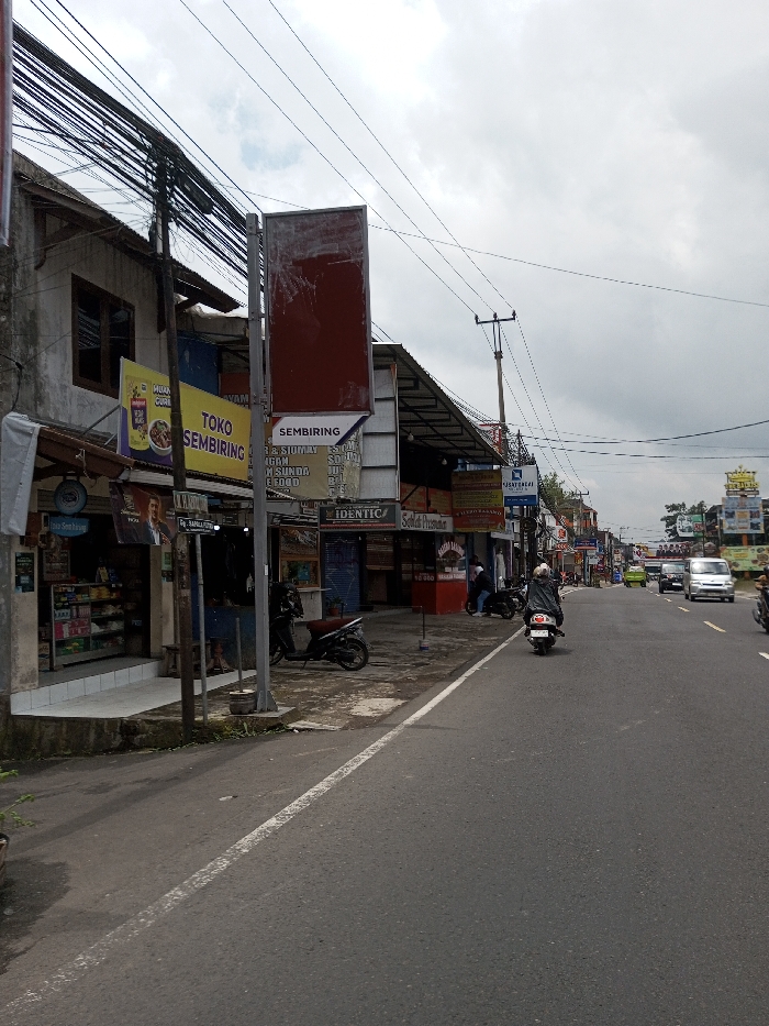 This image depicts a street scene in the city of Chendenbji, Bhutan, under an overcast sky. The view is from a paved roadside, looking down a two-lane asphalt road. On the left, a row of two-story commercial buildings lines the street, featuring various storefronts with awnings and signage.
Essential details include:
* **Environment:** The street features a grey asphalt surface with a faded white line marking the edge of the lane. A concrete curb separates the road from the adjacent sidewalk and buildings. Above, a dense network of black utility wires crisscrosses between multiple wooden and metal poles. The sky is largely grey and cloudy.
* **Buildings and Signage:** The buildings on the left exhibit light-colored facades, some with exposed brick or concrete textures, and corrugated metal awnings over storefronts. Prominent signs include a yellow sign for "TOKO SEMBIRING" and "HUANG CURRY" above a shop displaying packaged goods, a large rectangular red advertising panel on a pole, and other signs with text such as "SEMBIRING," "IDENTIC," and "Sekolah Primagama." One sign also features a depiction of a man's upper body.
* **Vehicles:** On the road, a person wearing a dark jacket and helmet rides a white scooter away from the camera in the right lane. Further down, a white SUV and other cars are visible, indicating traffic. Two dark-colored scooters are parked on the sidewalk on the left, near the storefronts. A partial view of another motorcycle is visible on the far right edge of the frame, moving away from the camera.
* **People:** Besides the scooter rider, at least two individuals are visible near the parked scooters and storefronts on the left side of the street, one possibly seated.
* **Objects:** A dark utility pole laden with wires stands prominently on the left sidewalk. Some minimal greenery is visible at the base of a pole on the far left.