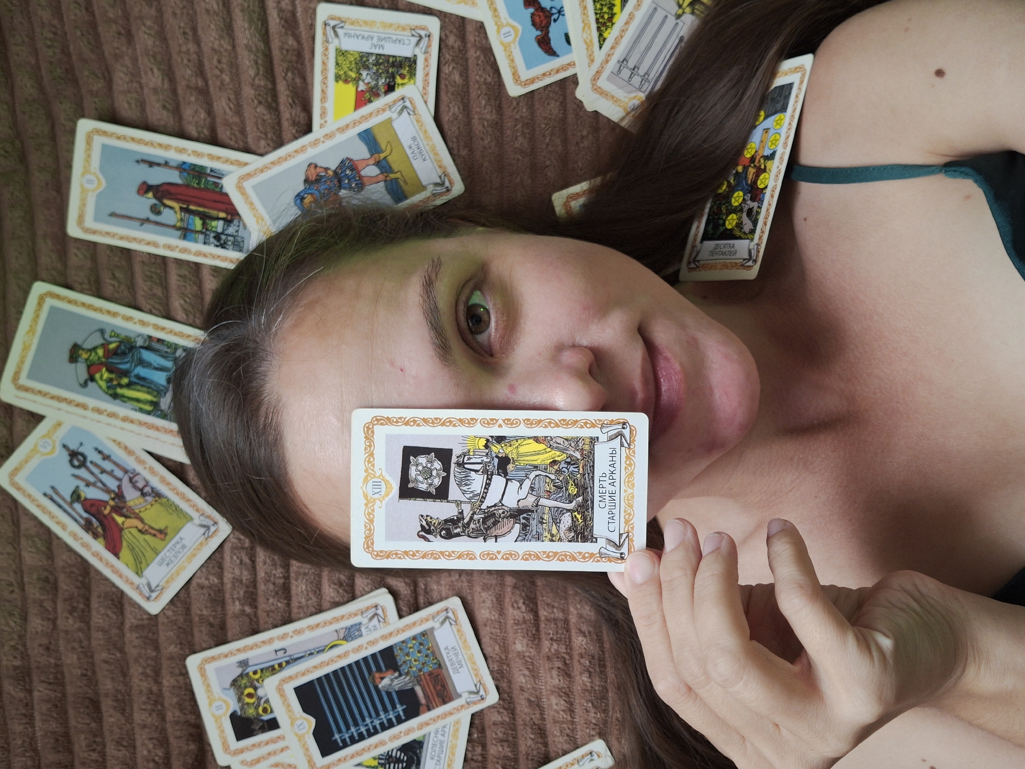 A person is lying on a brown, textured surface, with numerous tarot cards scattered around their head. One tarot card, identified as "Death, Major Arcana," is held in front of the person's face, obscuring their left eye and partially their nose. The card depicts a skeleton on horseback carrying a flag with a rose emblem, with figures and a landscape in the background. The person has long brown hair and is wearing a dark green strap. Other visible cards include "Six of Wands," "Two of Wands," "Nine of Swords," "Page of Diamonds," and "Ten of Pentacles." The setting appears to be an indoor environment. There is no discernible connection to the city of Chaikovsky, Russia.