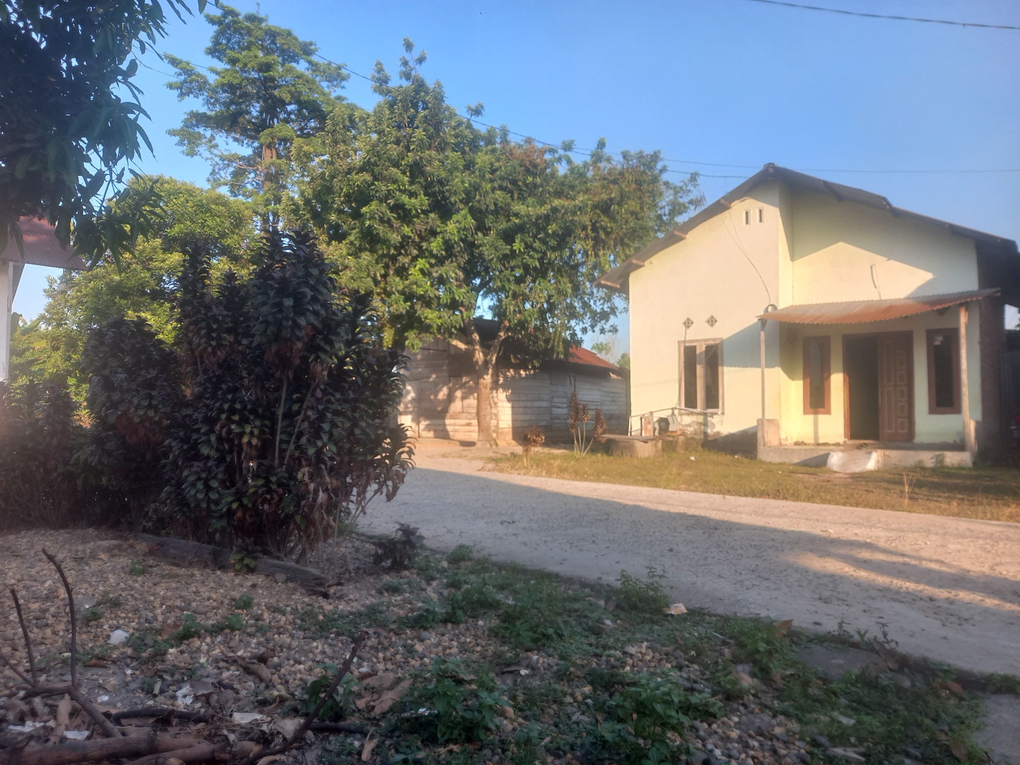 The image depicts a rural scene in Kasang Pudak, Indonesia. A dirt road runs diagonally across the frame, separating a foreground of rocky ground with sparse vegetation and scattered debris from the background. To the right of the road is a light-colored, single-story house with a corrugated metal awning over the entrance and several windows. Adjacent to this house, to the left and further back, is a smaller, weathered wooden structure. Lush green trees and dense foliage are prominent throughout the scene, with a large tree casting a shadow on the road. The sky is clear and blue, suggesting it is daytime, likely morning or late afternoon due to the lighting. There are no people visible in the image. The overall impression is one of a simple, tropical residential area.