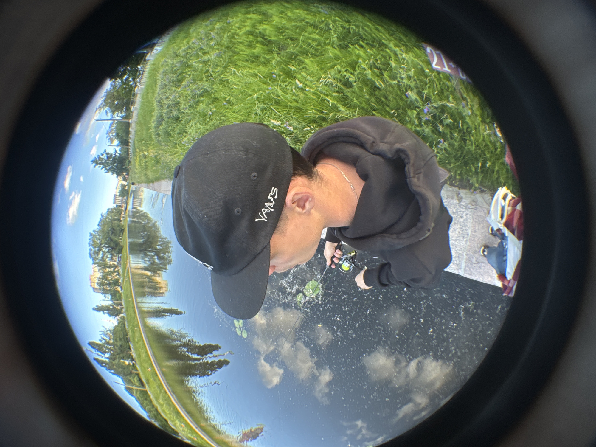 In Saint Petersburg, Russia, a young person wearing a black hoodie and a black baseball cap with white "Vans" branding is fishing in a canal. The fishhook is in the water, and lily pads are visible. The canal reflects the sky, trees, and a building. The surrounding area is grassy with trees and a path. The image is captured with a fisheye lens, giving it a wide, distorted perspective.
