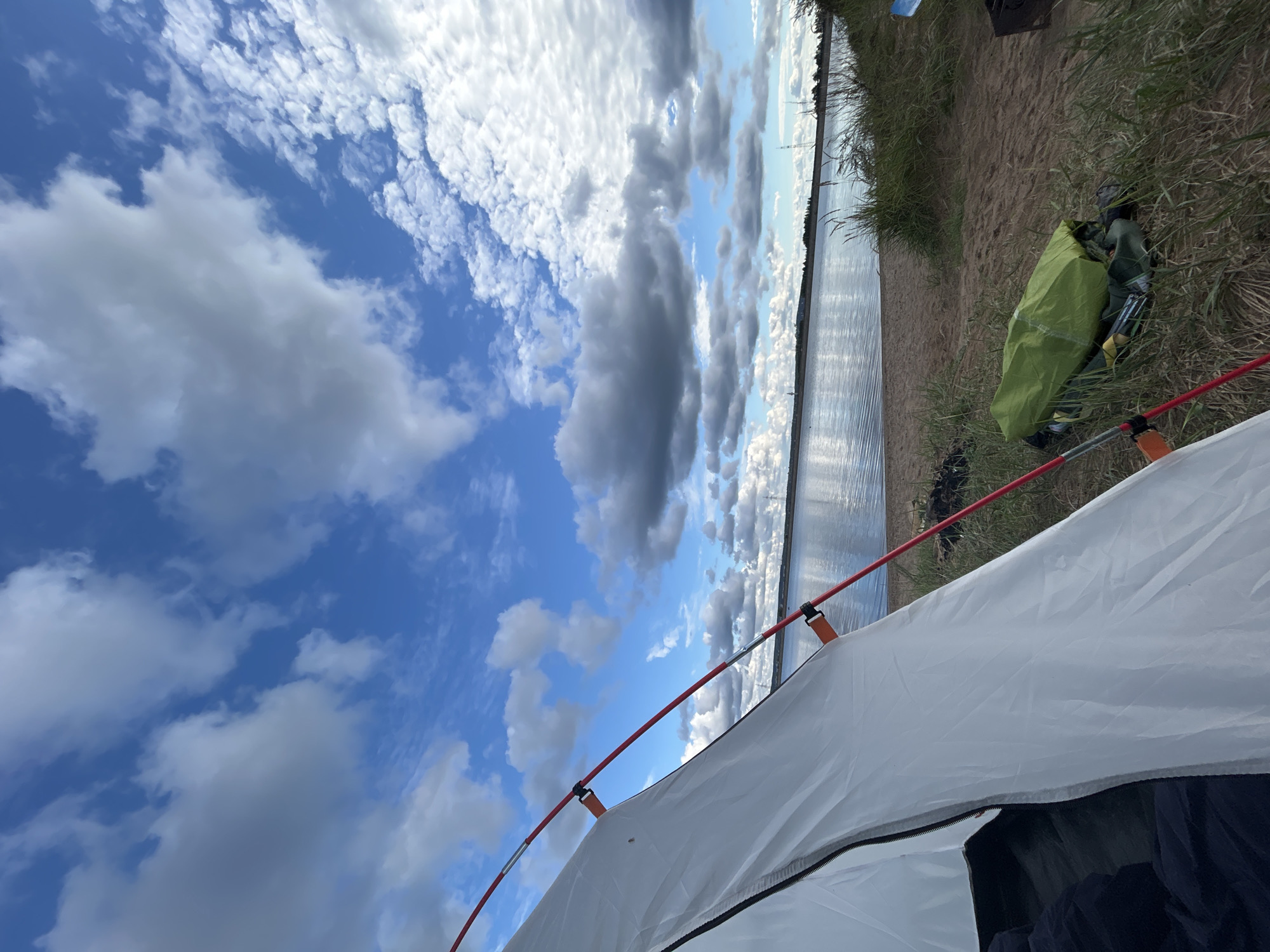 A campsite is set up on a sandy beach beside a calm body of water under a cloudy blue sky. In the foreground, the white fabric of a tent is visible, with red poles extending outwards. Next to the tent, a green sleeping bag or pack rests on the sand amongst tall, dry grass. The water stretches out to the horizon, where a faint treeline and several distant structures, possibly power lines or industrial buildings, can be seen. The sky is filled with a mixture of white and gray clouds, with patches of blue peeking through. The overall scene suggests an outdoor recreational setting.