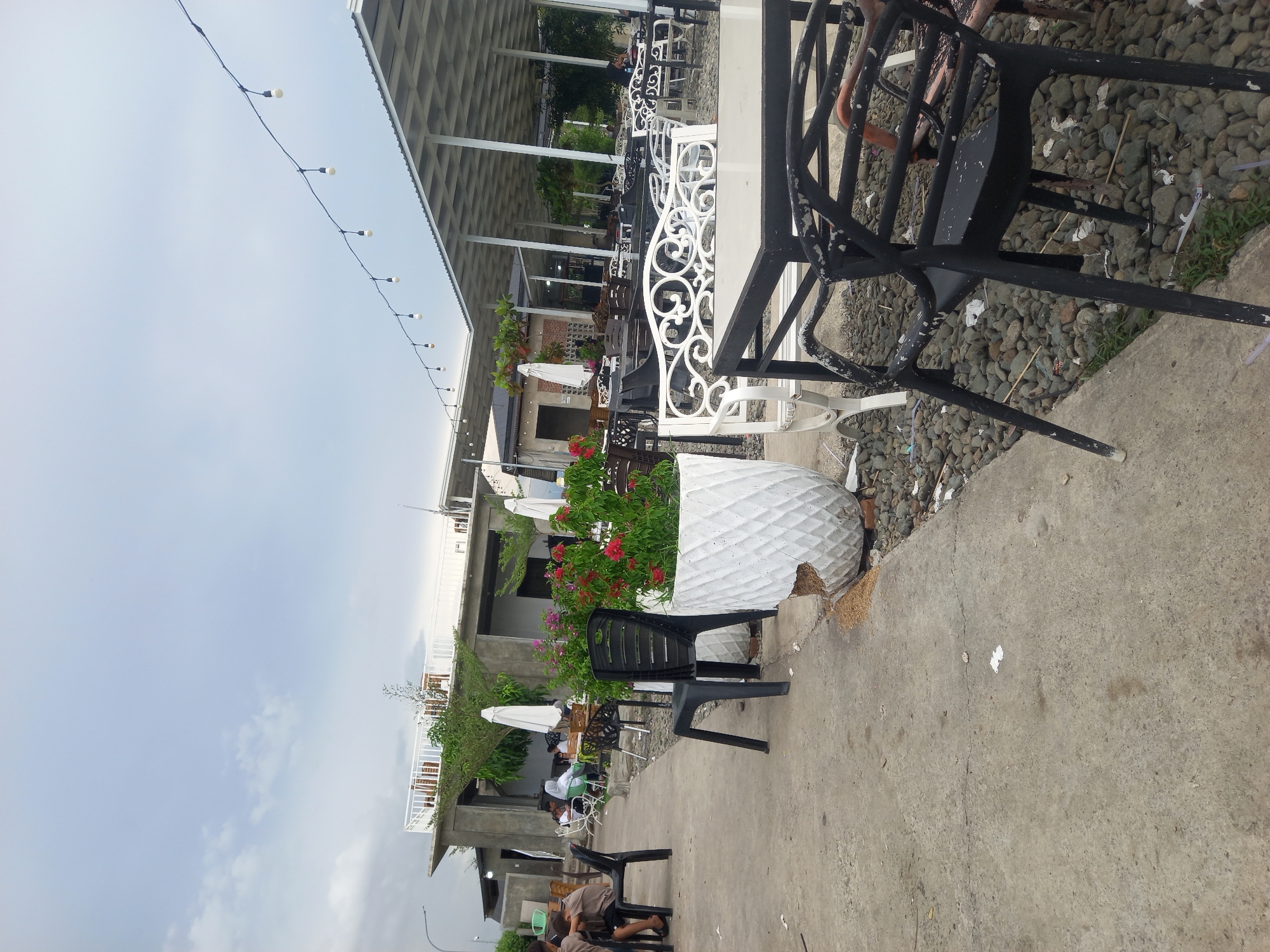 The image depicts an outdoor seating area at a cafe or restaurant in Cotmalem, Indonesia. The scene features multiple tables and chairs arranged on a concrete patio, with some areas covered by white awnings supported by pillars. Several people are visible, some seated at tables and others walking. In the foreground, there are black plastic chairs and a white decorative bench. Large white planters with red flowers are placed along the pathway. The ground is a mix of concrete and gravel. String lights are hung overhead. In the background, there is a multi-story building with a white railing on an upper level, surrounded by greenery. The sky is overcast.