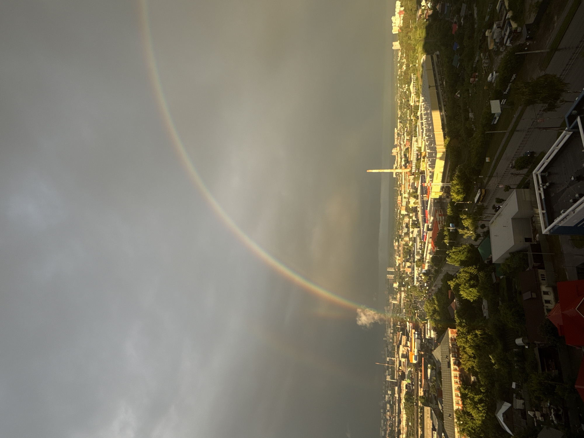 This is an aerial, wide-angle shot of Izhevsk, Russia, captured under a dramatic, overcast sky. The city stretches out below, a mix of industrial buildings, residential areas, and dense green trees. A prominent feature is the double rainbow arching across the sky, with the primary rainbow brighter and more defined than the secondary one. The rainbow appears to originate from a plume of white smoke or steam rising from an industrial area.
The cityscape includes several large, light-colored industrial buildings, one of which is illuminated by the sun breaking through the clouds. There's a tall, slender smokestack visible, and various other buildings of different sizes and architectural styles. Roads are visible, with some vehicles, including a yellow bus, on them, suggesting ongoing activity. The foreground of the image shows rooftops of residential buildings, some with red roofs, and an abundance of trees and greenery, indicating a city with pockets of nature. The overall atmosphere is one of a post-rain scene, with a sense of a city emerging from a storm, illuminated by the ethereal glow of a rainbow. The lighting suggests it is either late afternoon or early morning, with the sun low on the horizon.