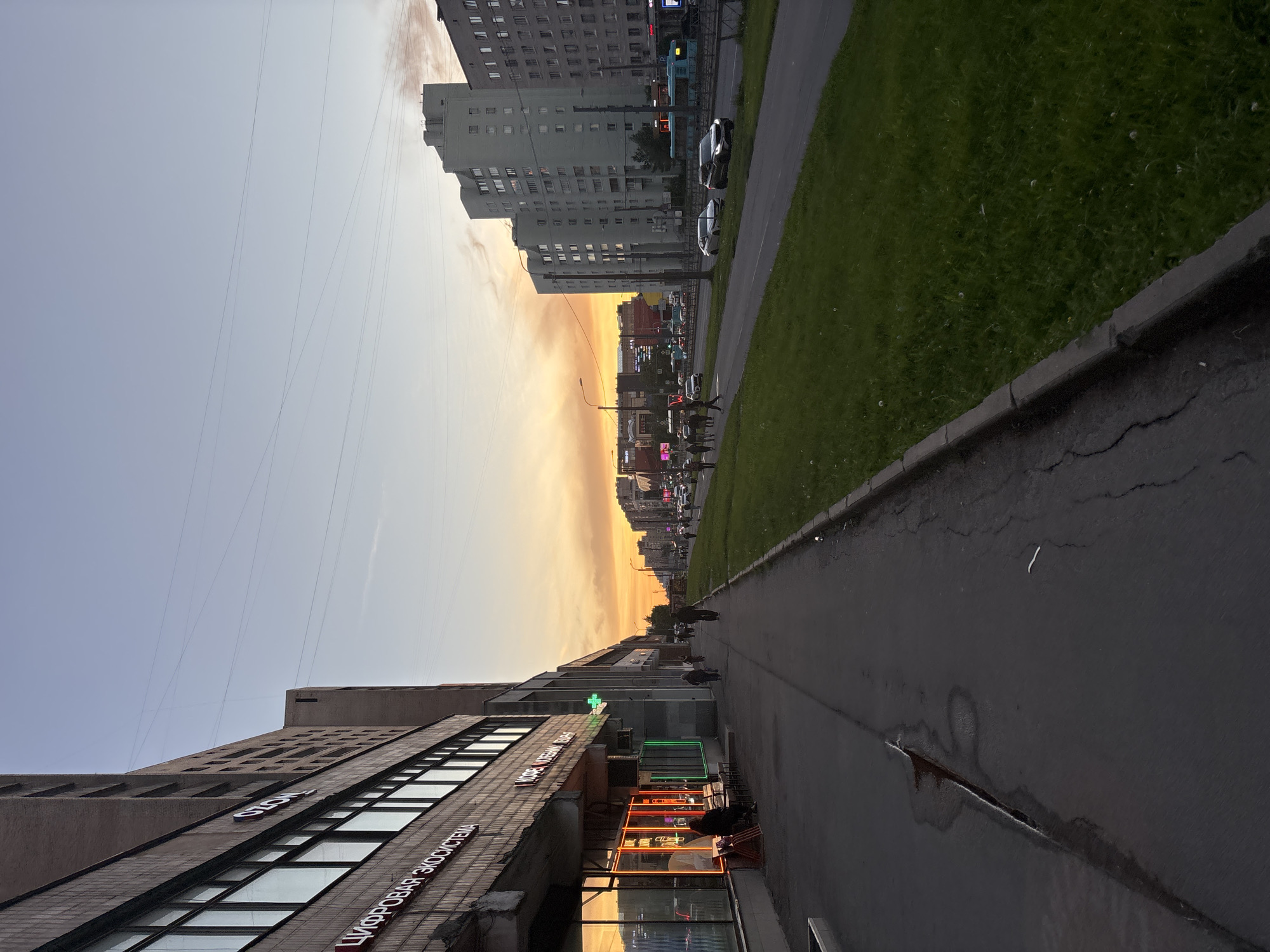 The image depicts a street scene at dusk in Sinyavino, Russia, with a dramatic sunset casting an orange glow across the sky. On the left side of the street, a multi-story building with large windows is visible, featuring neon signage for "OZON" and "ЦИФРОВАЯ ЭКОСИСТЕМА". A sidewalk runs along the building, with a few people walking and a bench outside a store. The sidewalk appears wet in places. Across the street, a road with several lanes of traffic is lined with residential buildings and green grassy areas. Vehicles, including cars and a bus, are visible on the road. Overhead power lines crisscross the sky. The overall impression is a typical urban evening scene in a Russian city.