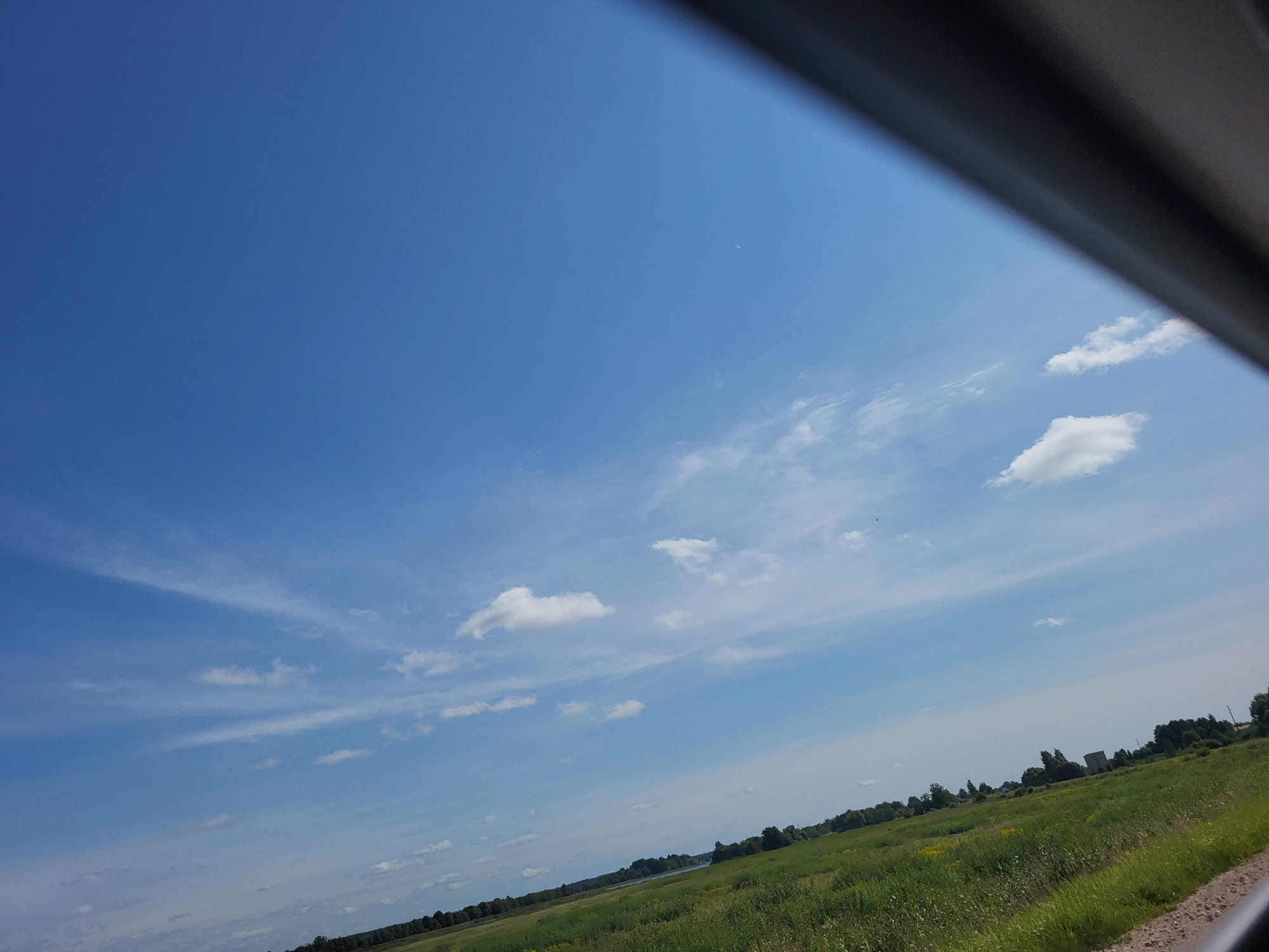 The image depicts a rural landscape under a clear blue sky with scattered white clouds. A diagonal dark object, possibly a vehicle roof or window frame, intrudes from the top right corner, obscuring a portion of the sky. The lower half of the image shows a vast green field, likely agricultural land, with some yellow wildflowers visible. A line of trees borders the field in the distance, and beyond that, a body of water, possibly a river or lake, is discernible. Several scattered buildings and structures are visible among the trees, suggesting a small settlement. The overall impression is one of a serene, open countryside. The location is identified as Kalnciems, Latvia.