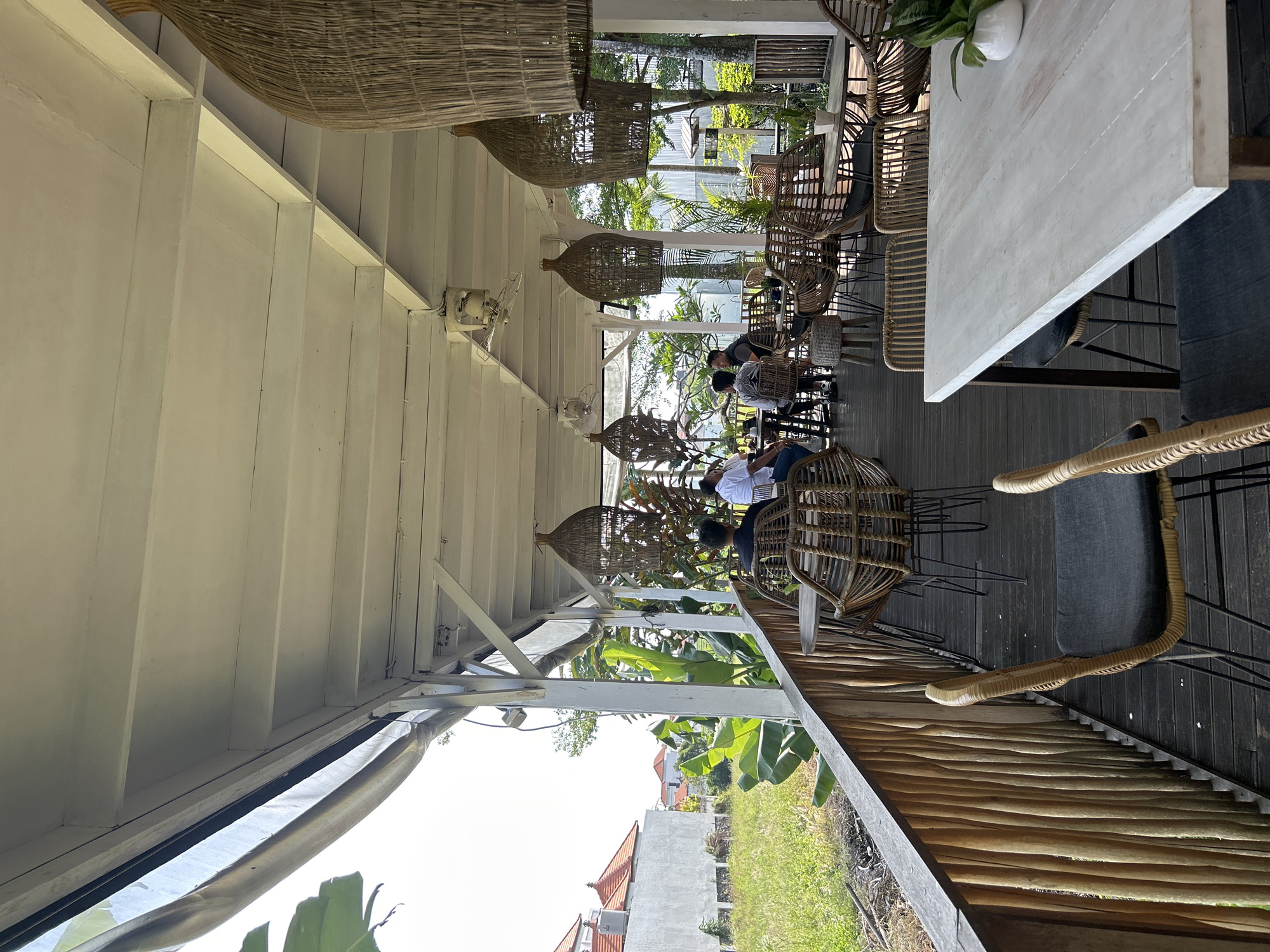 The image depicts an outdoor seating area of a cafe in Mengwi, Indonesia, bathed in bright daylight. The cafe features a wooden deck floor and a white, open-air roof structure supported by white pillars. Several tables and chairs, made of woven rattan and metal, are arranged on the deck. Large, woven pendant lamps hang from the ceiling. In the background, lush green vegetation and trees are visible, alongside a white building with orange-tiled roofs. Some patrons are seated at tables, engaged in conversation or looking at their phones. The overall ambiance suggests a relaxed, tropical setting.