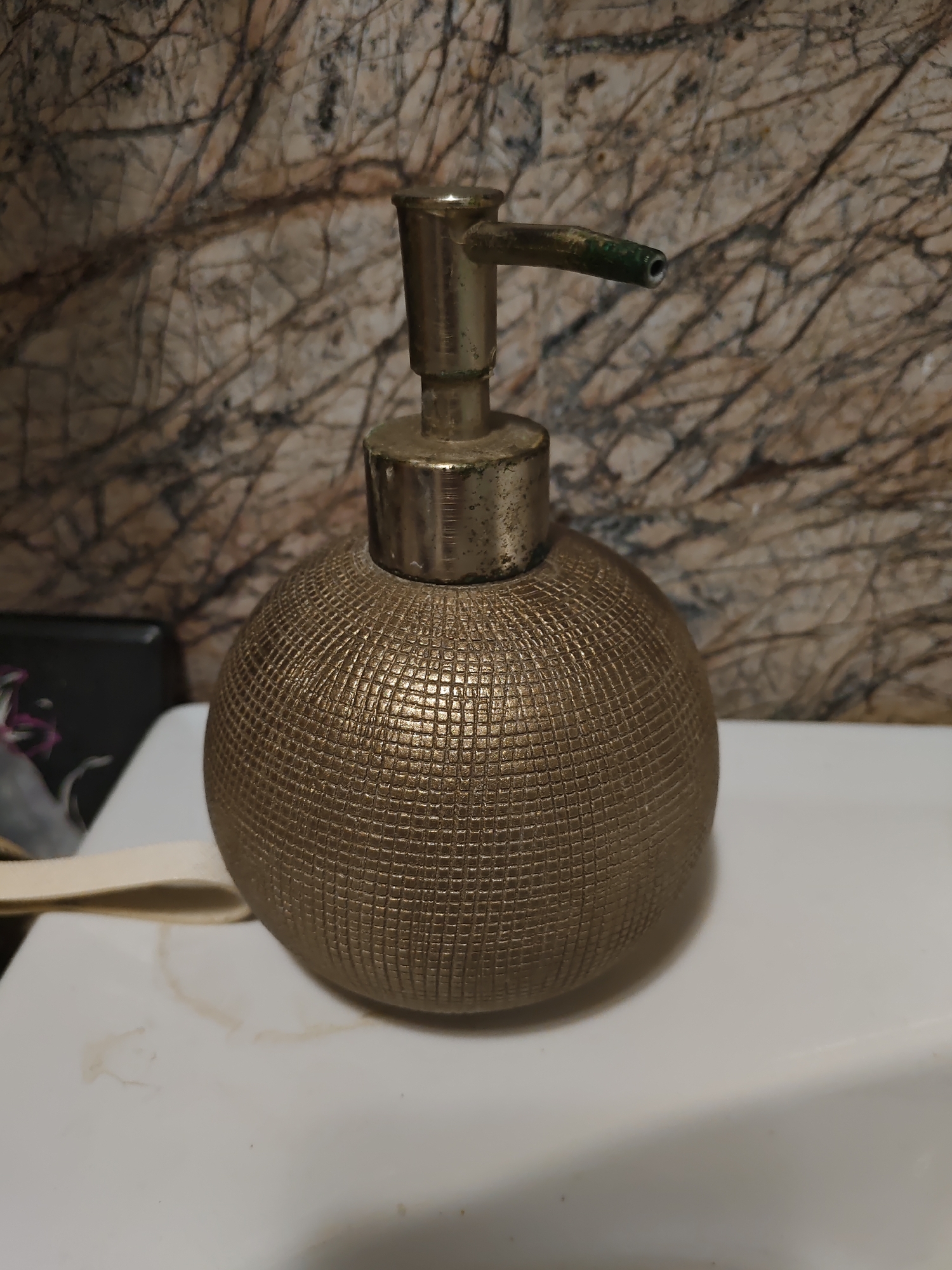 The image shows a close-up, eye-level, static shot of a decorative, spherical, golden dispenser with a textured surface. The dispenser has a metallic pump mechanism with a green-tipped nozzle. It is placed on a white, smooth surface, possibly a sink or countertop. Behind the dispenser, a textured, marble-like wall is visible. To the left of the dispenser, a dark object with some pinkish-purple floral or abstract design is partially visible, along with a light-colored strap or loop. The lighting appears to be from the front, casting subtle shadows. The image provides no information or context related to the city of Rostov-on-Don, Russia.