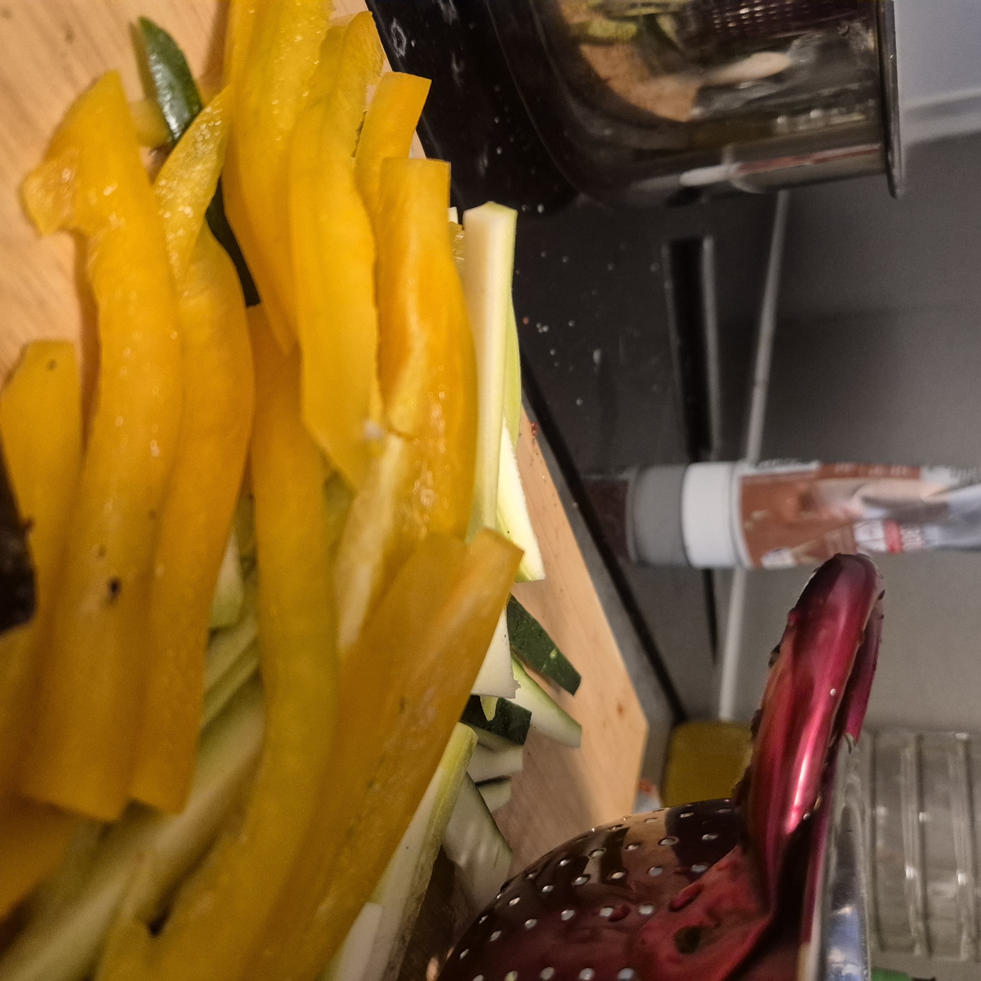The image shows a close-up of cut yellow bell peppers and zucchini on a wooden cutting board. Beside the cutting board are cooking utensils, including a red colander and what appears to be a portion of a pan. A tube is also partially visible, sitting on what seems to be a kitchen counter. Based on the context, the scene is likely located in a kitchen somewhere in Strasbourg, France.