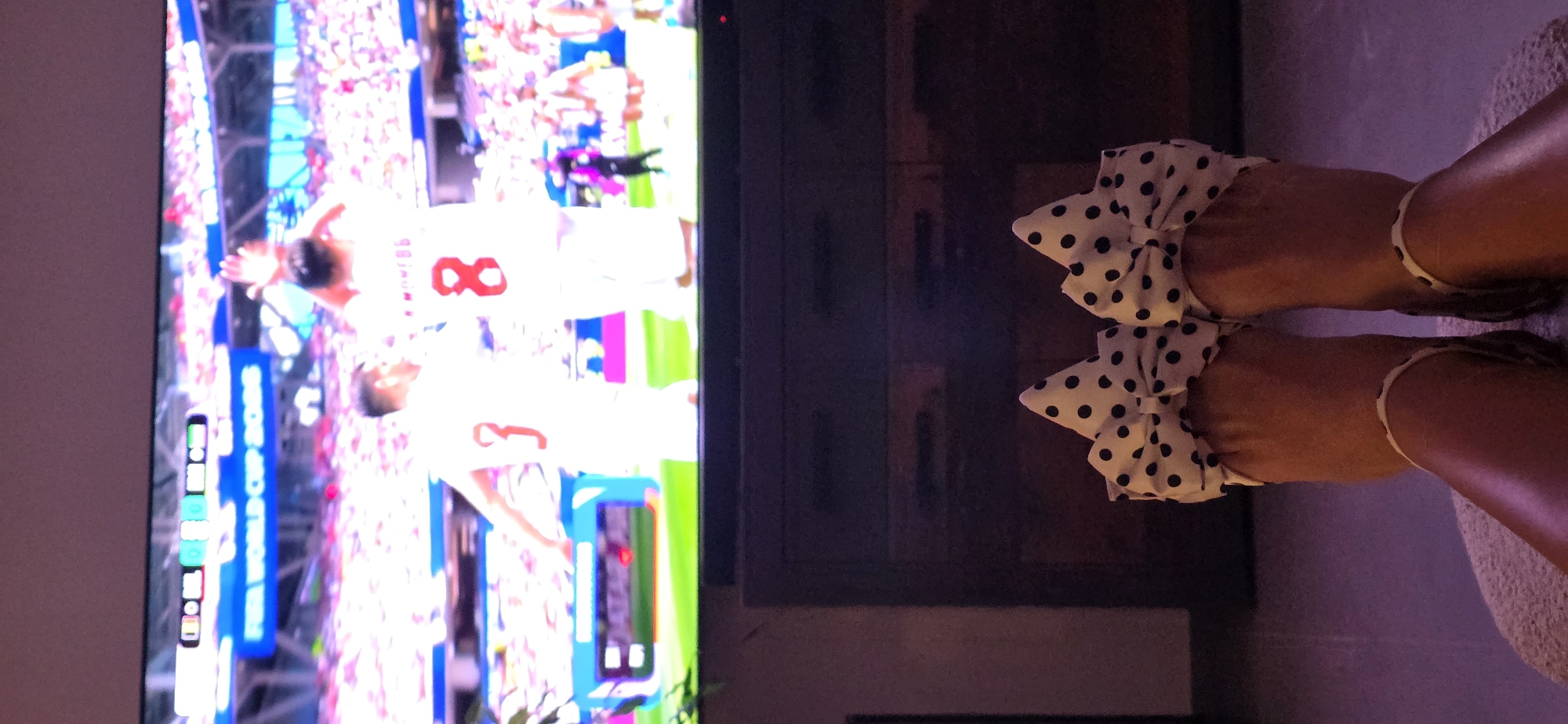 The image captures a person's feet wearing polka dot slippers with bows, resting on a textured surface, in front of a television. The television screen displays a soccer match, possibly from the FIFA World Cup 2018, with players in white jerseys and a scoreline indicating 0-0. The background of the television screen shows a stadium filled with spectators. The overall scene suggests a relaxed atmosphere, perhaps at home, watching a sporting event. There is no discernible information about the city of Lieli, Switzerland, in this image.