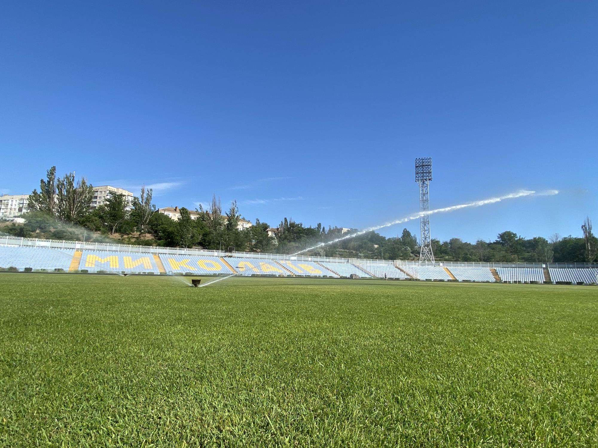 An eye-level, wide shot captures a sunlit stadium on a clear day. The foreground is dominated by a lush green, well-maintained grass field, with a sprinkler system actively irrigating the turf in the mid-ground. In the background, tiered stadium seating, predominantly blue with yellow lettering that appears to spell "MMKO," curves around the field. A tall floodlight tower stands on the right side of the image, with water spraying from a nozzle high up on its structure, creating a white mist against the blue sky. Trees and buildings are visible on a hillside beyond the stadium seating, indicating an urban or semi-urban environment. The sky is a vibrant, cloudless blue.