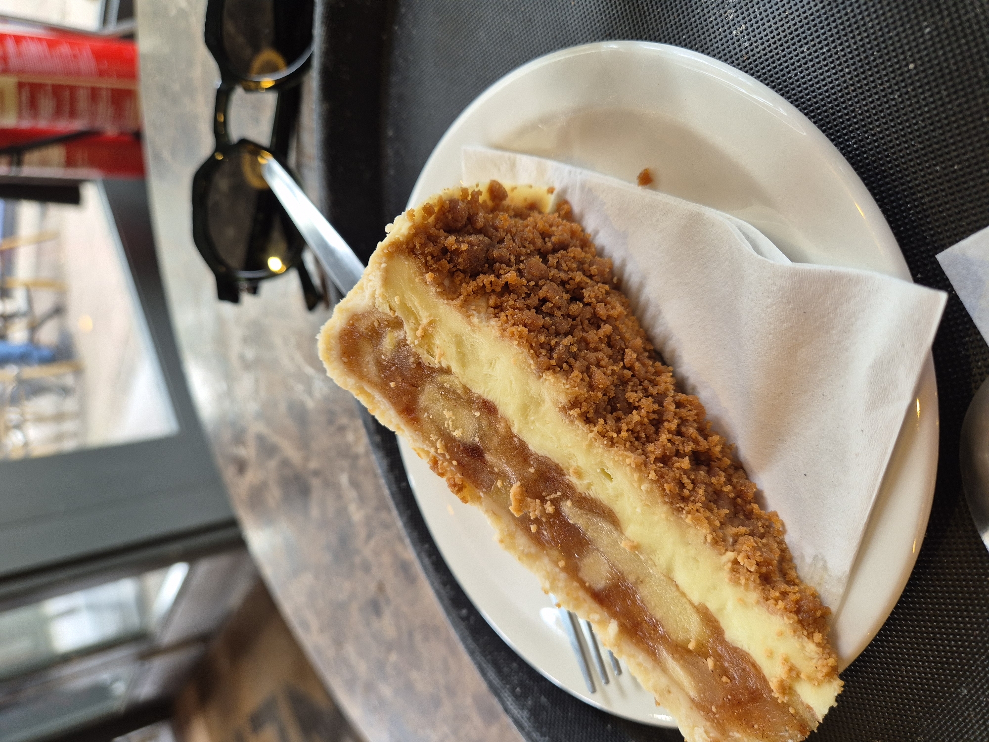 A slice of dessert is prominently featured on a white ceramic plate, captured from a slightly overhead perspective. The dessert is a layered cheesecake, possibly an apple crumble cheesecake, with a creamy yellow layer, a lighter fruit filling (likely apple), and a generous topping of brown crumble. A white paper napkin is partially tucked under the dessert, and a silver fork is visible beneath the front of the slice.
To the upper left of the plate, a pair of black sunglasses with dark lenses and a silver hinge detail rests on a dark surface. The plate and sunglasses are positioned on a dark, textured mat, which itself sits on a lighter, possibly marbled table surface. The background is softly blurred, revealing hints of an outdoor or window view to the left, including a red object (possibly a beverage can) and other indistinct architectural shapes.
This casual dining scene, likely in a cafe or restaurant in West Suffolk, United Kingdom, is well-lit, suggesting daytime. No people or legible text are visible.