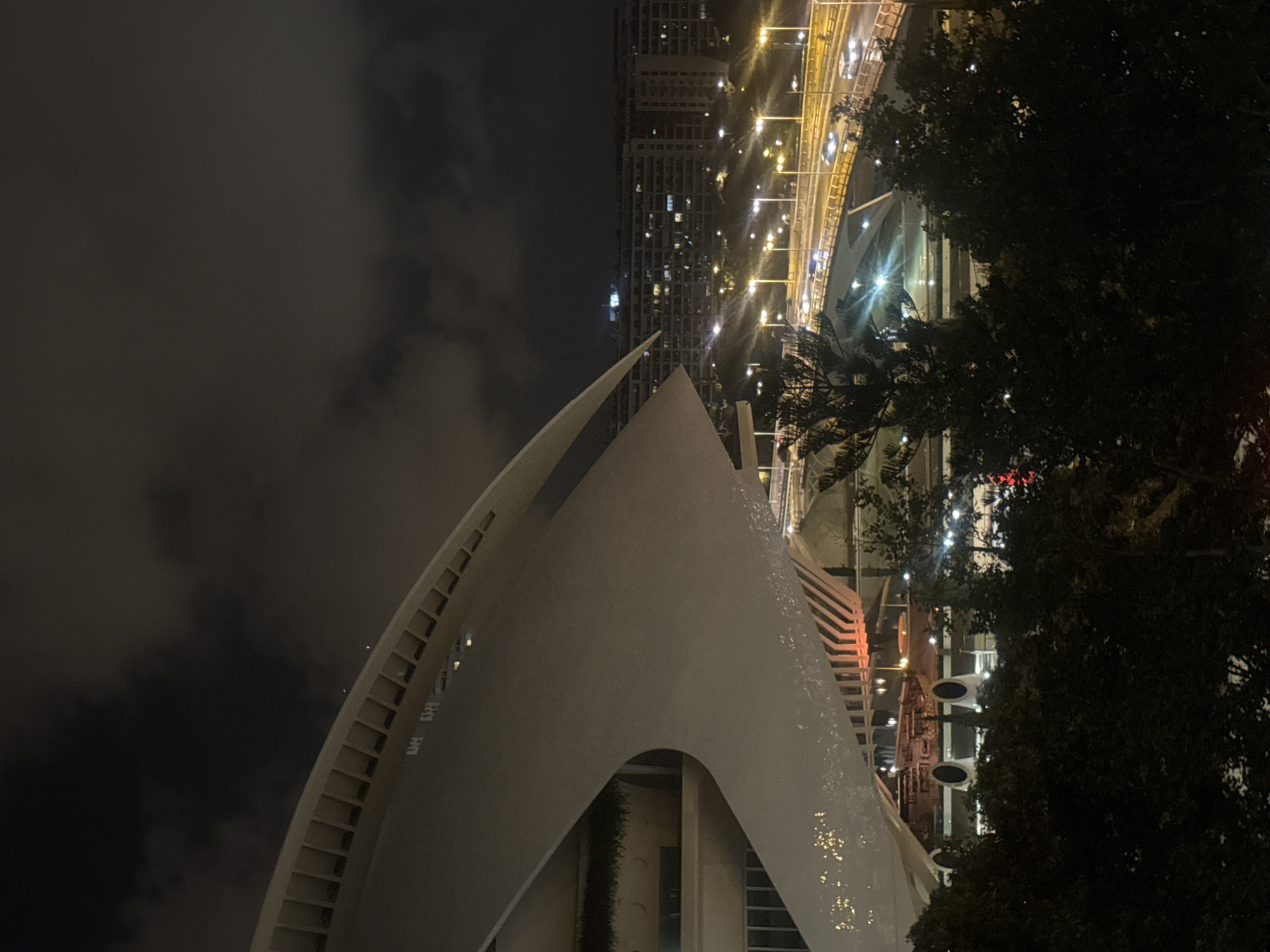 The image captures a nighttime view of the City of Arts and Sciences in Valencia, Spain, specifically focusing on the L'Hemisfèric building. The foreground is dominated by the distinctive white, curved structure of the building, its modern architecture illuminated from below by warm orange lights. Beyond the L'Hemisfèric, a multi-lane highway curves through the scene, with cars visible as streaks of light due to the long exposure. Streetlights line the highway, casting a yellow glow. In the background, a dense urban landscape of apartment buildings is visible, their windows lit, creating a pattern of lights against the dark sky. The sky itself is cloudy, obscuring the stars and contributing to the overall low-light ambiance. There are no people visible in the frame.