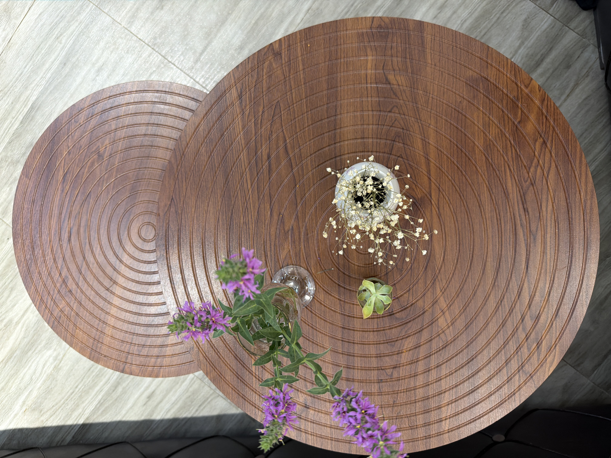 A top-down view shows two round, wooden coffee tables with concentric circle carvings on their surfaces. The tables are positioned slightly offset, with the upper table partially covering the lower one. On the larger, lower table, there are three decorative items: a clear glass vase with purple flowers and green leaves, a small succulent plant in a round container, and a white pot filled with dried white flowers. The tables are placed on a light-colored, wood-patterned floor. The lighting suggests a well-lit indoor environment, possibly a living room or lounge area. There are no people visible in the image. The scene appears to be a static display of decorative objects.